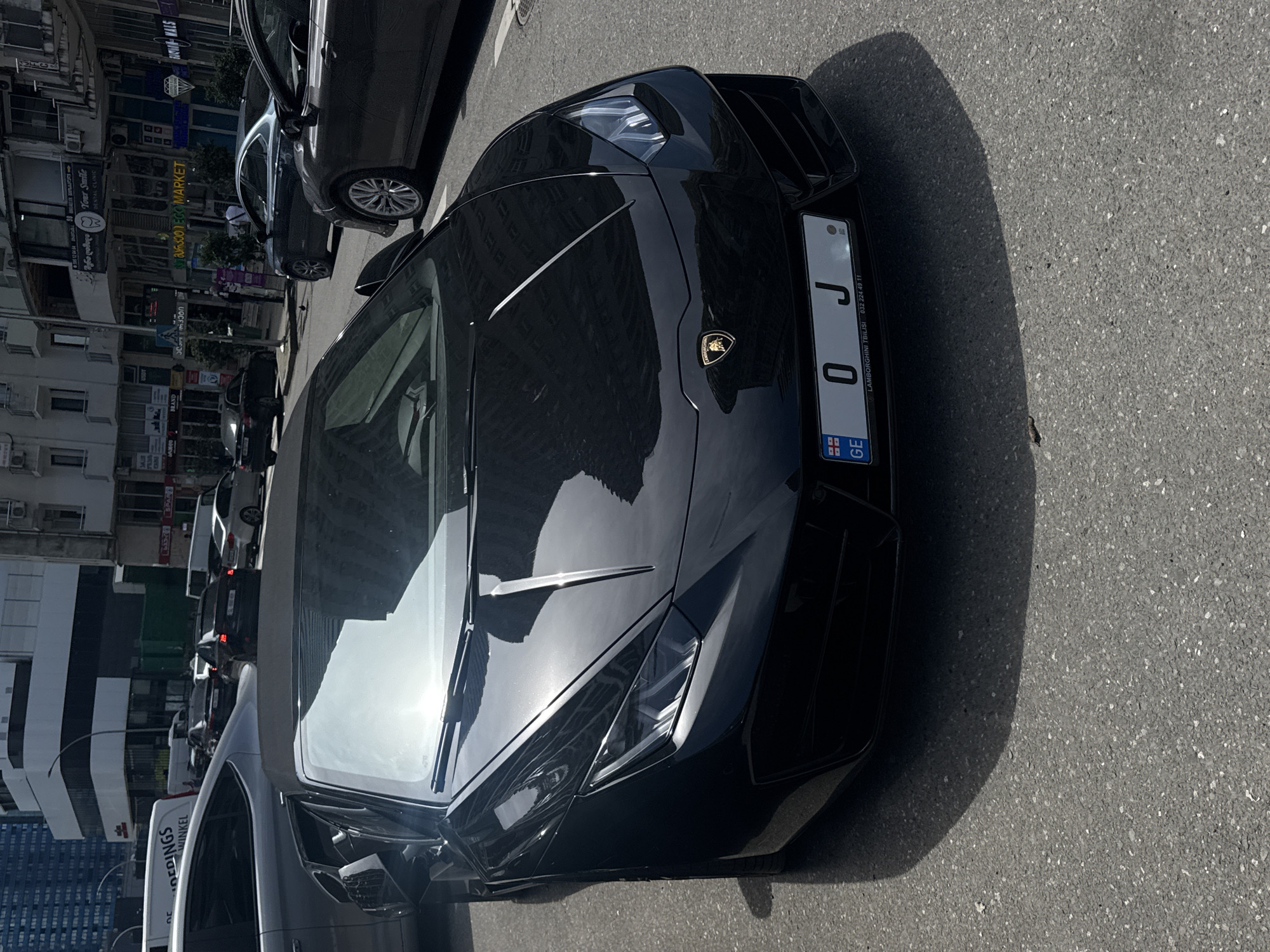 A high-angle, eye-level shot of a black Lamborghini Huracan Spyder parked on a city street. The car is the primary subject, with a Georgian license plate reading "GE 0J". The Lamborghini logo is visible on the hood. Sunlight reflects off the car's surface.
Behind the Lamborghini, other parked vehicles are visible. On the left, buildings line the street, with signs in both Georgian and English, indicating a business district. Signboards are visible.
The location is likely in Batumi, Georgia, based on the text on the car license plate. The weather seems sunny. The scene is a typical urban landscape with a luxury vehicle as a focal point.