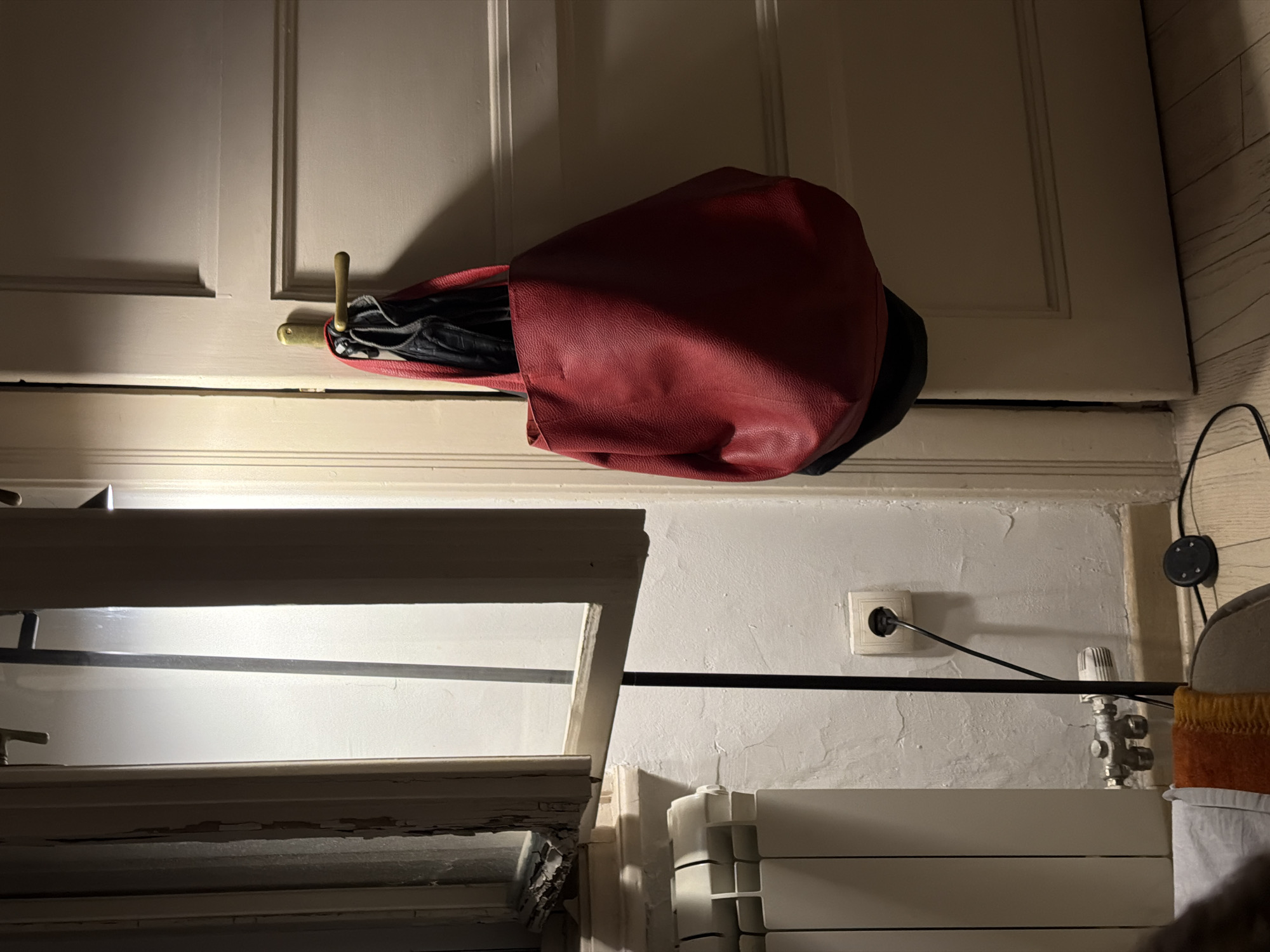 An interior scene, likely a hallway or room in an apartment in Vracar Urban Municipality, Serbia. A red leather tote bag and a black jacket are hanging from a brass door handle on a closed, light-colored door. To the left, a white radiator is visible below a partially open window with weathered wooden frames and a frosted glass pane. A thin, black lamp post extends upwards near the window. On the wall behind the lamp post, a white electrical outlet with a black plug and cord is visible, along with plumbing fixtures near the floor. The floor appears to be made of light-colored wood planks. A dark circular object, possibly a smart speaker, with a black cord is on the floor in the lower right corner.