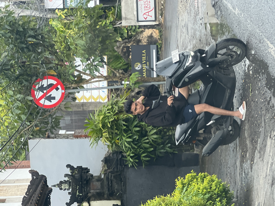 A young man sits on a black scooter parked on a wet street. He wears a black hoodie, navy shorts, and white flip-flops. His left hand rests on his head. Behind him is a lush green bush and a stone structure with traditional Balinese carvings. In the background, a "no passing" sign is visible amidst the trees, along with signs for "Mani Villa" and "Airis."