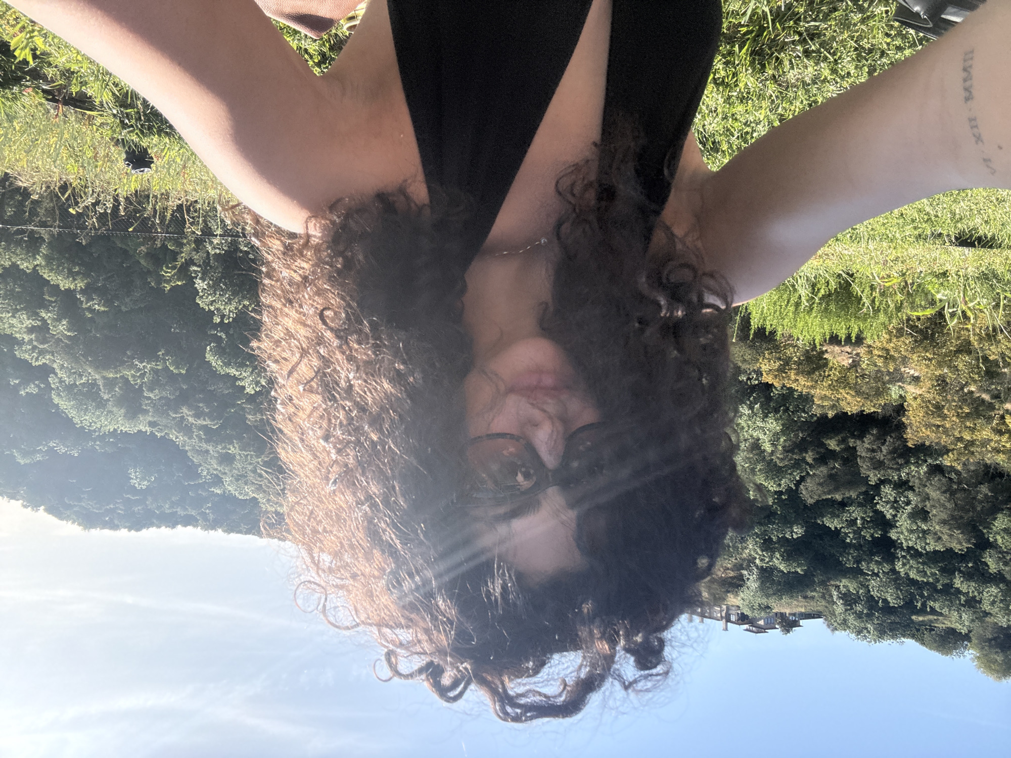 A person wearing sunglasses and a dark top is holding their arms up while taking a selfie outside on a sunny day. The background shows greenery, including trees and grass. There is also a hint of a building and the sky. The location is in Los Angeles, United States of America.