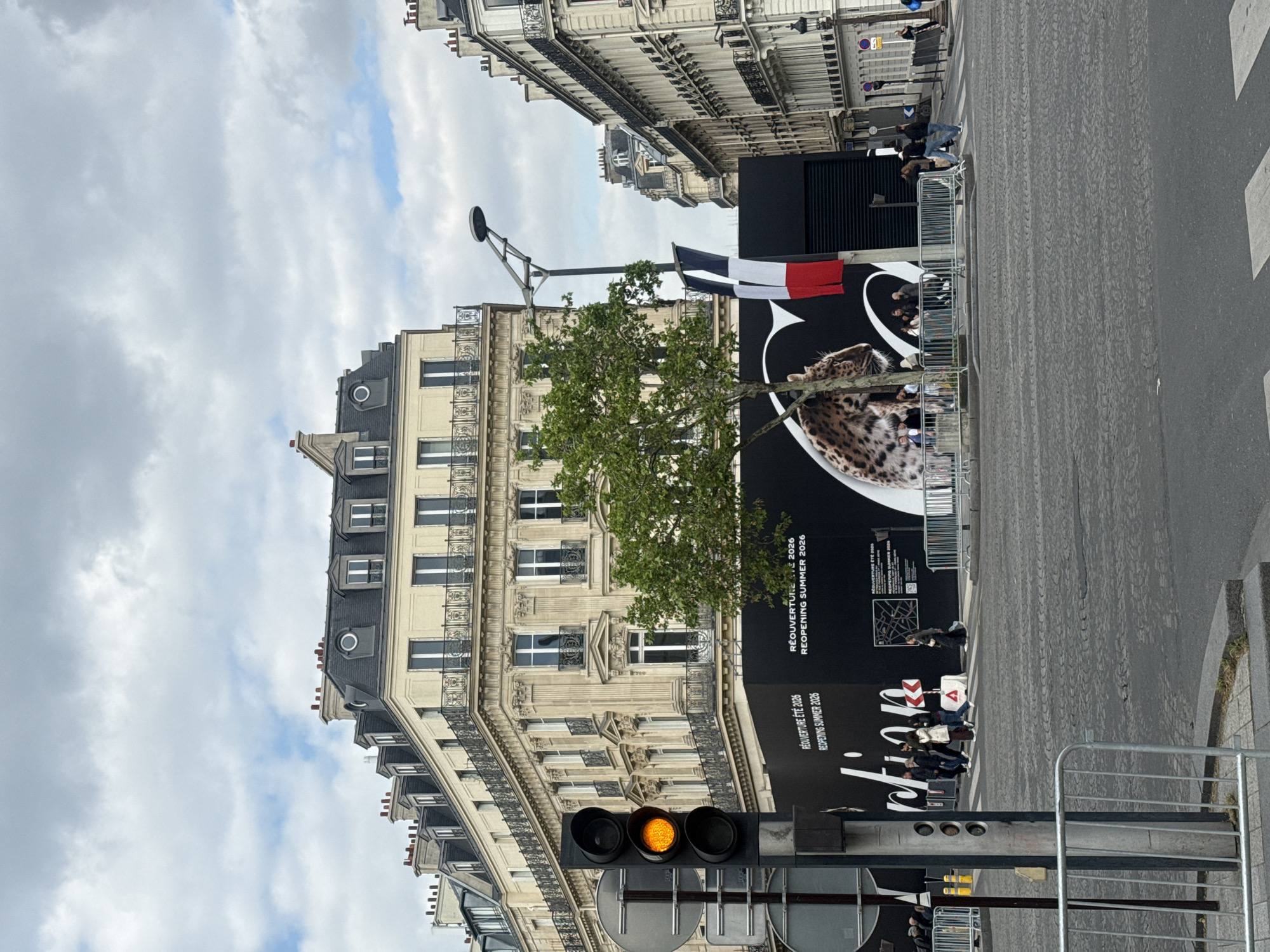 The image shows a street scene in Paris, France. A large black advertisement banner is prominently displayed, featuring a cheetah-like image with text including "REOUVERTURE ETE 2026 REOPENING SUMMER 2026." The French flag is attached to the banner. A tree is partially obstructing the view of the banner. Buildings with classic Parisian architecture are visible in the background, constructed of light-colored stone, with multiple stories and windows. A traffic light with a yellow light is in the foreground. Pedestrians are standing and sitting near the banner. The street is paved, and metal barriers are present. The sky is cloudy.