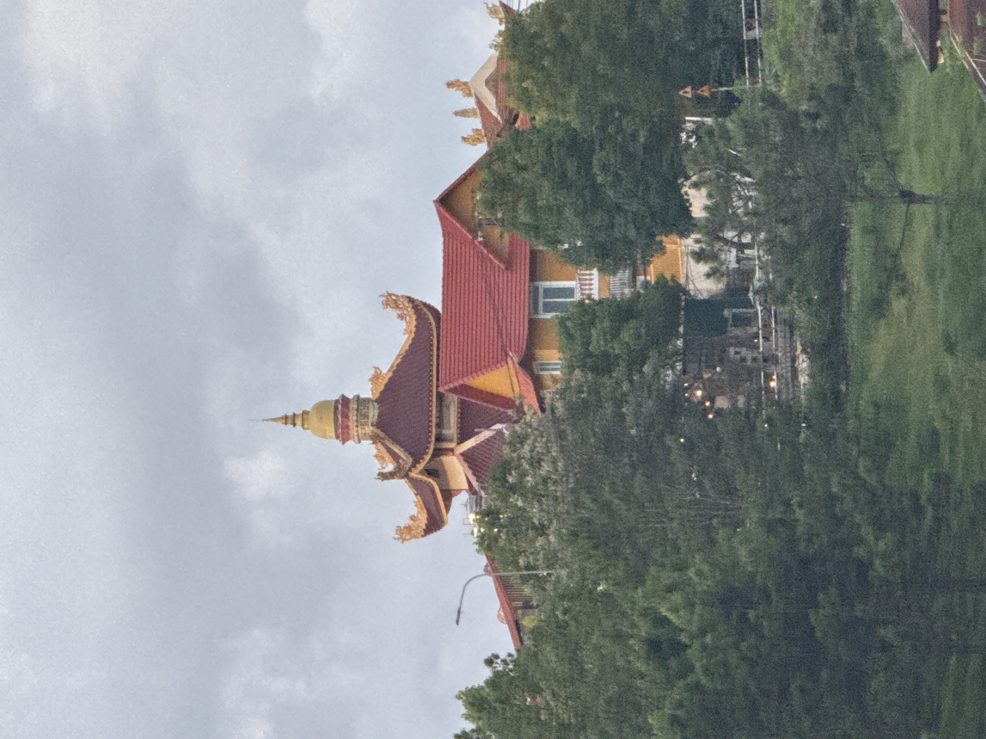 The image depicts a Buddhist pagoda, likely located on a hillside in Da Lat, Vietnam, given the architectural style. The pagoda features a prominent golden dome and intricate red and gold roof decorations with curved eaves. It is partially obscured by lush green trees that surround the structure, with some taller pine trees in the foreground. The sky is overcast and grey, suggesting a cloudy or possibly rainy day. In the middle ground, a section of a yellow building with a red-tiled roof is visible, adjacent to the pagoda. There are also signs of human activity, with a street light visible near the pagoda and a hint of outdoor lighting or decorations within the trees. The overall scene conveys a sense of serene religious architecture nestled within a natural landscape.