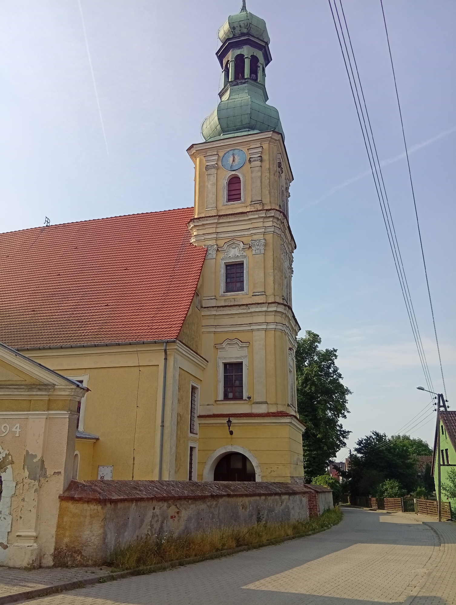 A low-angle, full outdoor shot shows a yellow, multi-story church building with a red tiled roof and a green domed steeple. The steeple has a clock with a blue face and red hands pointing to approximately 4:20. A stone wall with some overgrown weeds runs along the front of the church. A paved street with a cobblestone pattern leads away from the church, curving to the right. Power lines run diagonally across the sky. A green house with a wooden fence is visible on the right side of the street. The sky is clear and blue with a contrail from an airplane visible. The scene appears to be in a rural or semi-rural area. The church is located in Brzezia Łąka, Poland.