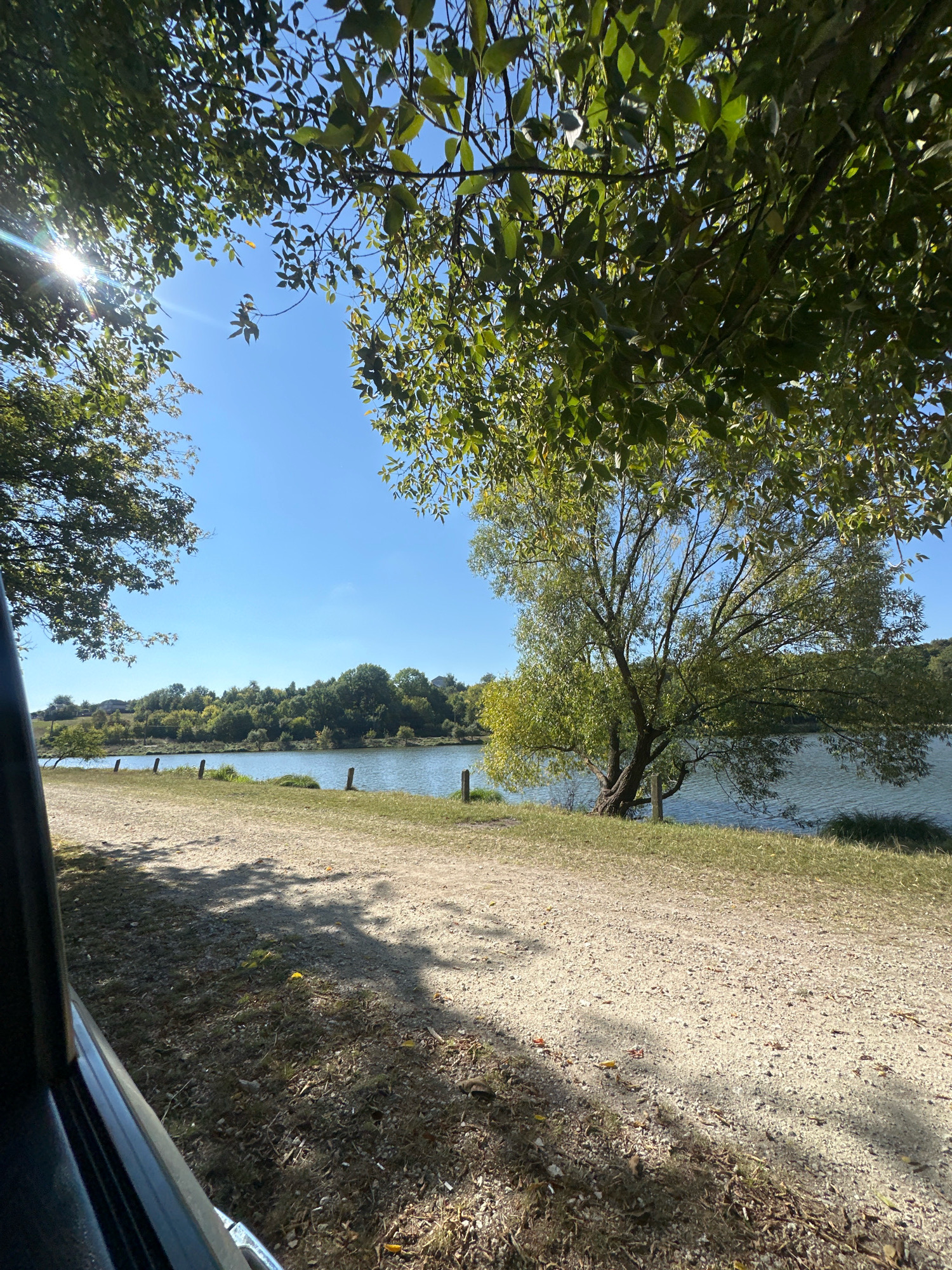 The image depicts a serene lakeside scene in Makiv, Ukraine, under a clear blue sky. The foreground is dominated by a dirt path, casting dappled shadows from overhead foliage. To the left, a dark vehicle is partially visible, with its edge extending into the frame. Beyond the path, a grassy bank slopes down to a calm body of water. Several wooden posts mark the transition from the grass to the water. On the far bank, a dense line of trees forms a verdant backdrop, with some buildings peeking through the foliage on a distant hill. A solitary, mature tree with sprawling branches stands near the water's edge, its leaves rustling gently. The sunlight glints through the leaves of trees in the upper left corner, creating lens flare. The overall atmosphere is one of natural beauty and tranquility.