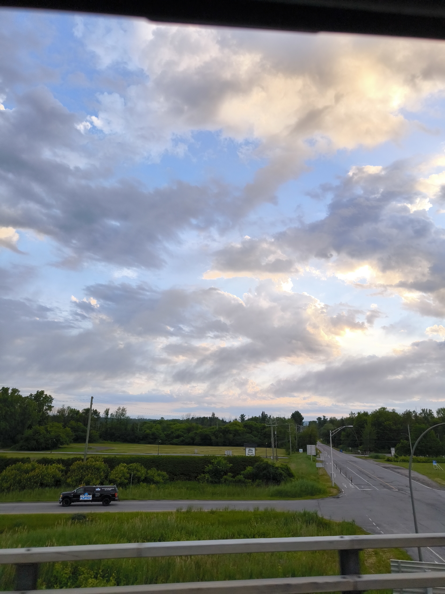 The image shows a highway scene in Saint-Lazare, Canada, with a black pickup truck driving on the road. The truck has "PUREY" written on its side, along with other decals. The road is a two-lane highway with grassy shoulders and trees lining the sides. The sky is cloudy with patches of blue and sunlight breaking through. In the background, there are more trees and some distant buildings. A railing is visible in the foreground, suggesting the photo was taken from an elevated position, possibly a bridge or overpass.