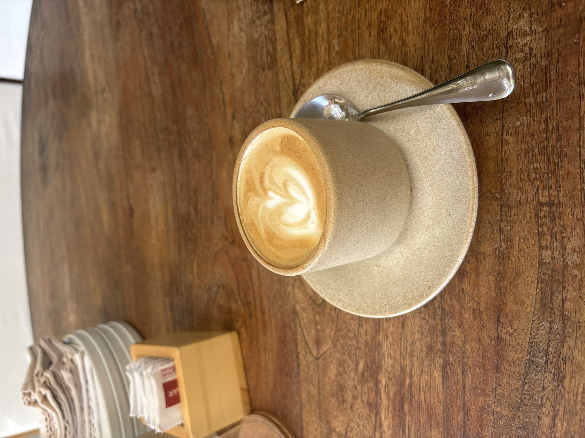 A tan ceramic coffee cup with latte art sits on a matching saucer, accompanied by a silver spoon. The cup and saucer are placed on a wooden table. Next to the cup are a stack of paper napkins and a small wooden container, presumably holding sugar packets. The setting suggests a cafe or restaurant, specifically located in Kuta Selatan, Indonesia, based on the image description.