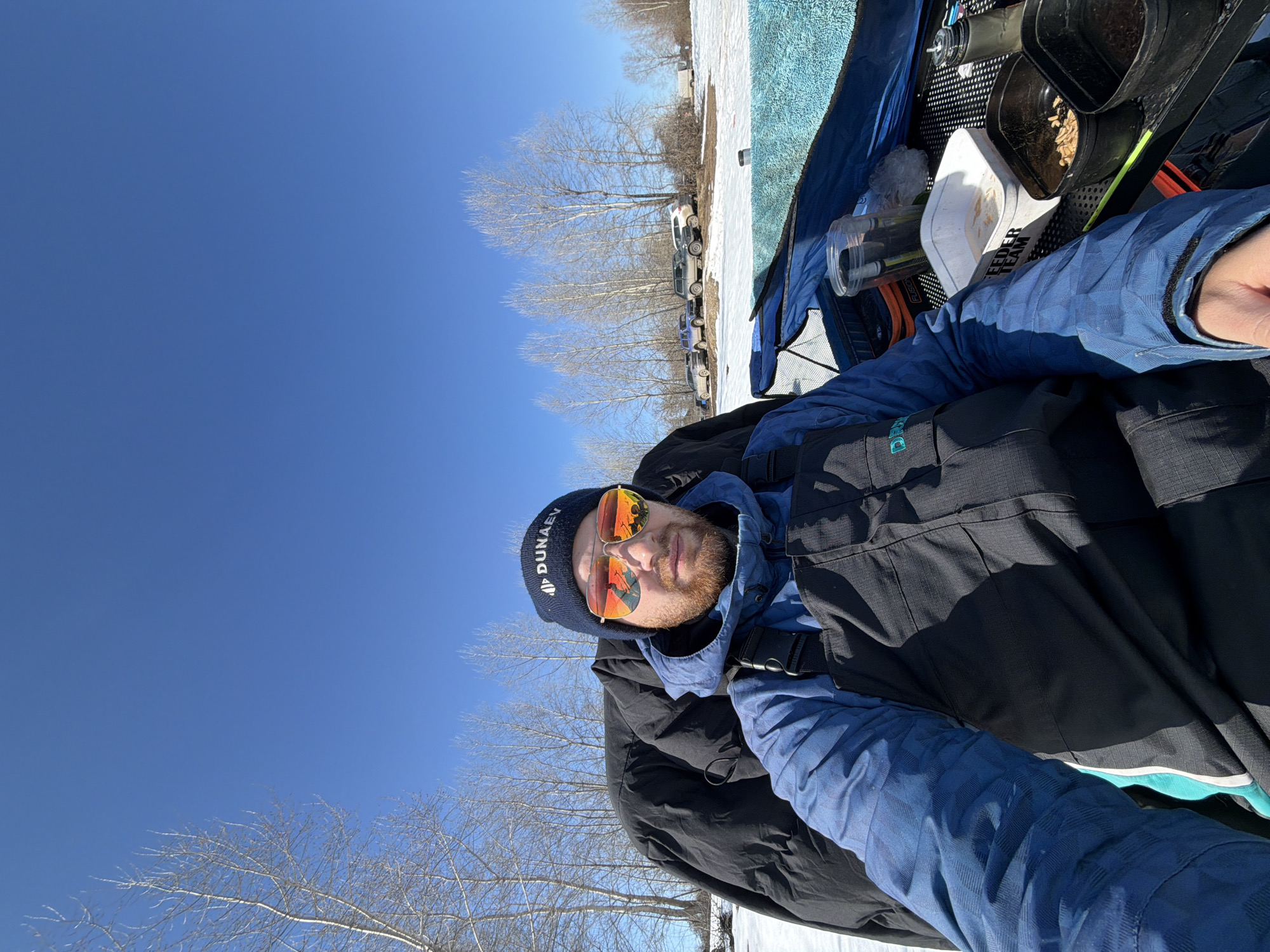 The image shows a man wearing a dark blue beanie with white lettering "DUNAEV," orange-tinted sunglasses, a blue patterned jacket, and a black vest. The man is presumably taking a selfie. Behind him, there's a blue tarp or tent, and several containers, some of which contain what appears to be fishing bait or pellets. In the background, there's snow on the ground, bare trees, and some parked cars. The sky is clear and blue. While the location of Noviy, Russia cannot be confirmed from the image, the overall scene suggests an outdoor setting during winter.