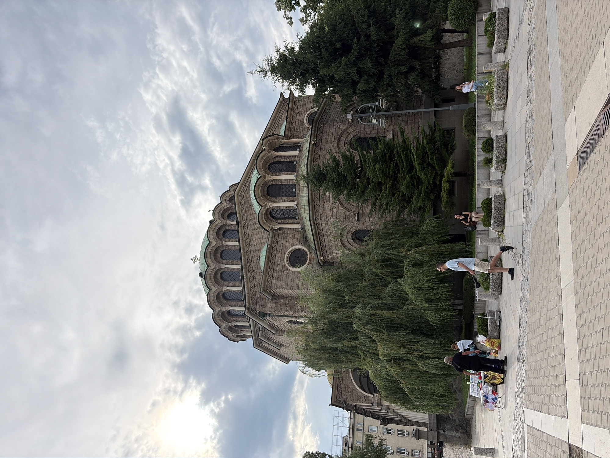 The image captures a view of the Alexander Nevsky Cathedral in Sofia, Bulgaria, under a cloudy sky with sunlight breaking through on the left. The massive stone cathedral, with its distinct green domes and arched windows, dominates the upper half of the frame. In the foreground, a paved plaza is visible, with several people present. To the left, a vendor with a cart displays items, and two individuals are seen interacting near it. A man in a light blue t-shirt and khaki shorts walks from left to right in the middle ground. Further back, a weeping willow tree and several fir trees are situated around the cathedral. Some people are seated on planters or benches on the right side of the plaza. The overall scene depicts a public space in front of a significant architectural landmark.