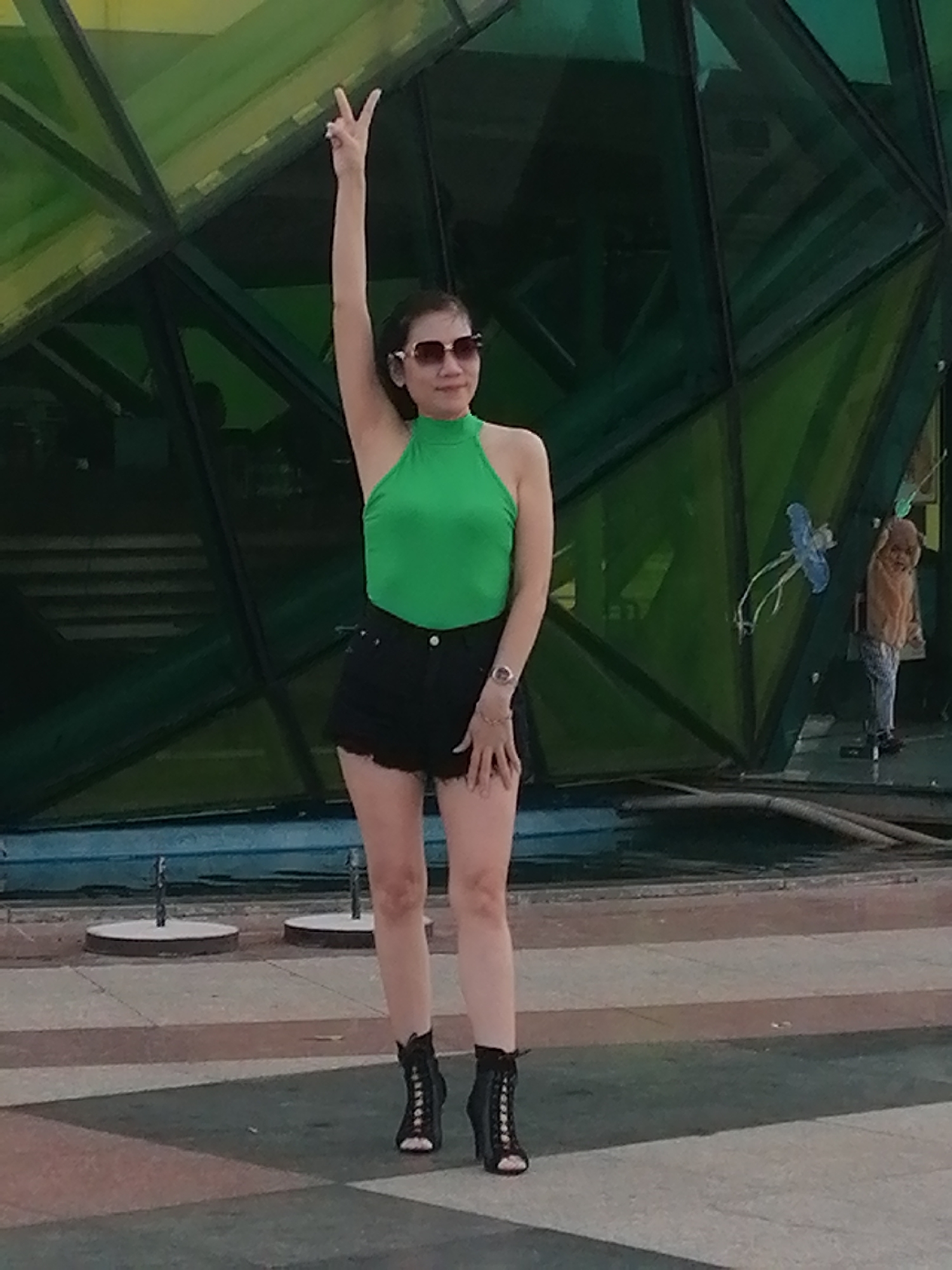 A woman stands in front of a geodesic dome-like structure with green, faceted panels. She is wearing a bright green halter top, black shorts, and black heeled boots. Her right arm is raised, with her index and middle fingers forming a peace sign. She is also wearing sunglasses. In the background, a person in a light brown hooded garment and grey pants is visible. The ground is paved with geometric patterns, and a body of water with fountains is present in the mid-ground. This scene is likely located at the Xuan Huong Lake area in Da Lat, Vietnam, known for its modern architecture and recreational spaces.