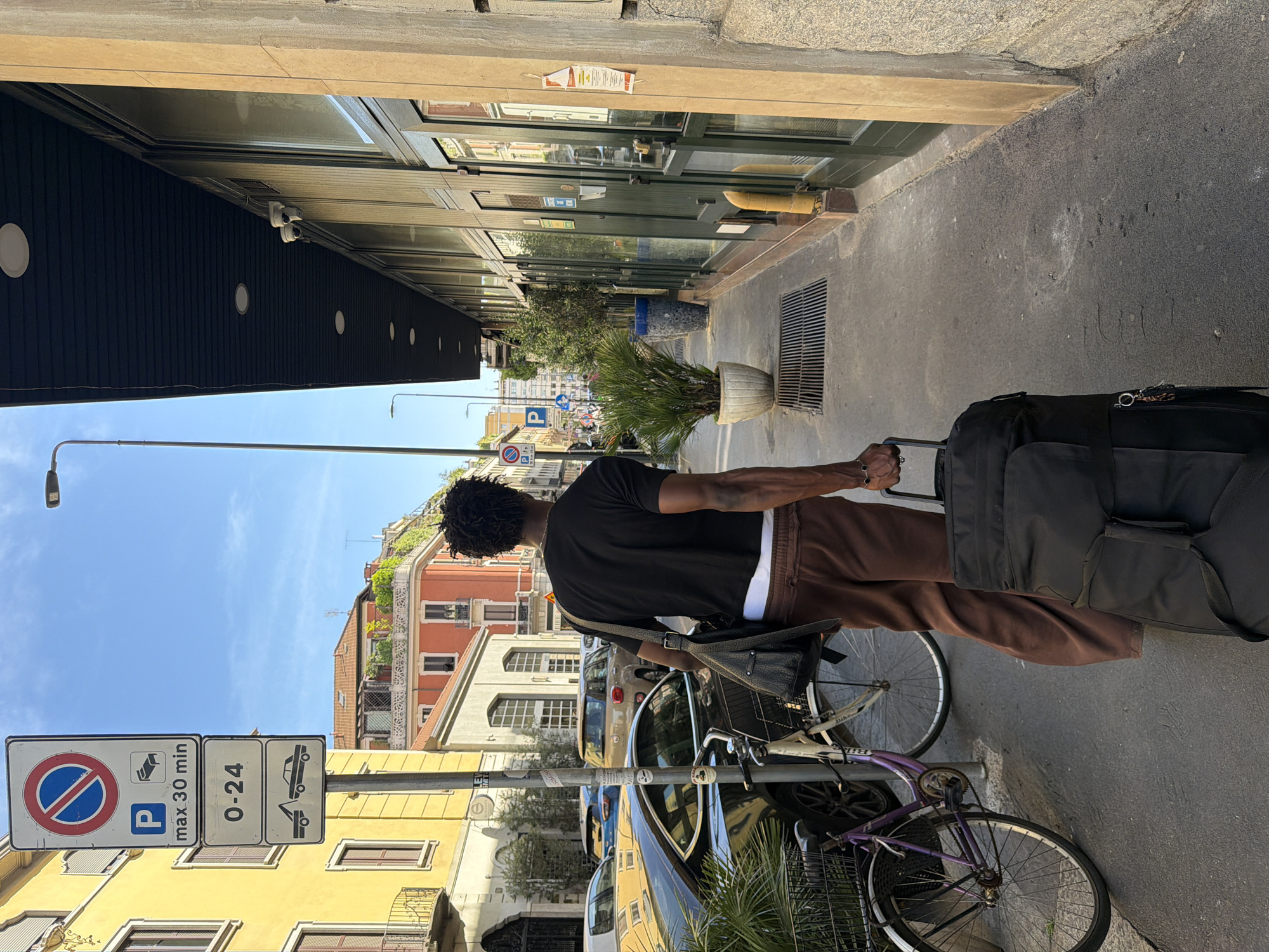 A person with dark, curly hair walks away from the camera down a sidewalk in Milan, Italy, pulling a large black suitcase. The person is wearing a black t-shirt and loose brown pants. A dark bag is slung over their left shoulder. They are walking on a paved sidewalk next to a street with parked cars, including a dark-colored sedan and a purple bicycle with a basket. Across the street, there are yellow and orange buildings with windows and balconies. A traffic signpost with parking regulations is visible to the left of the person. The street appears to be lined with shops or businesses with large glass doors and windows. Two security cameras are mounted on the exterior wall of one of the buildings, above the storefront. Potted plants are placed outside some of the establishments. The sky is clear and blue.