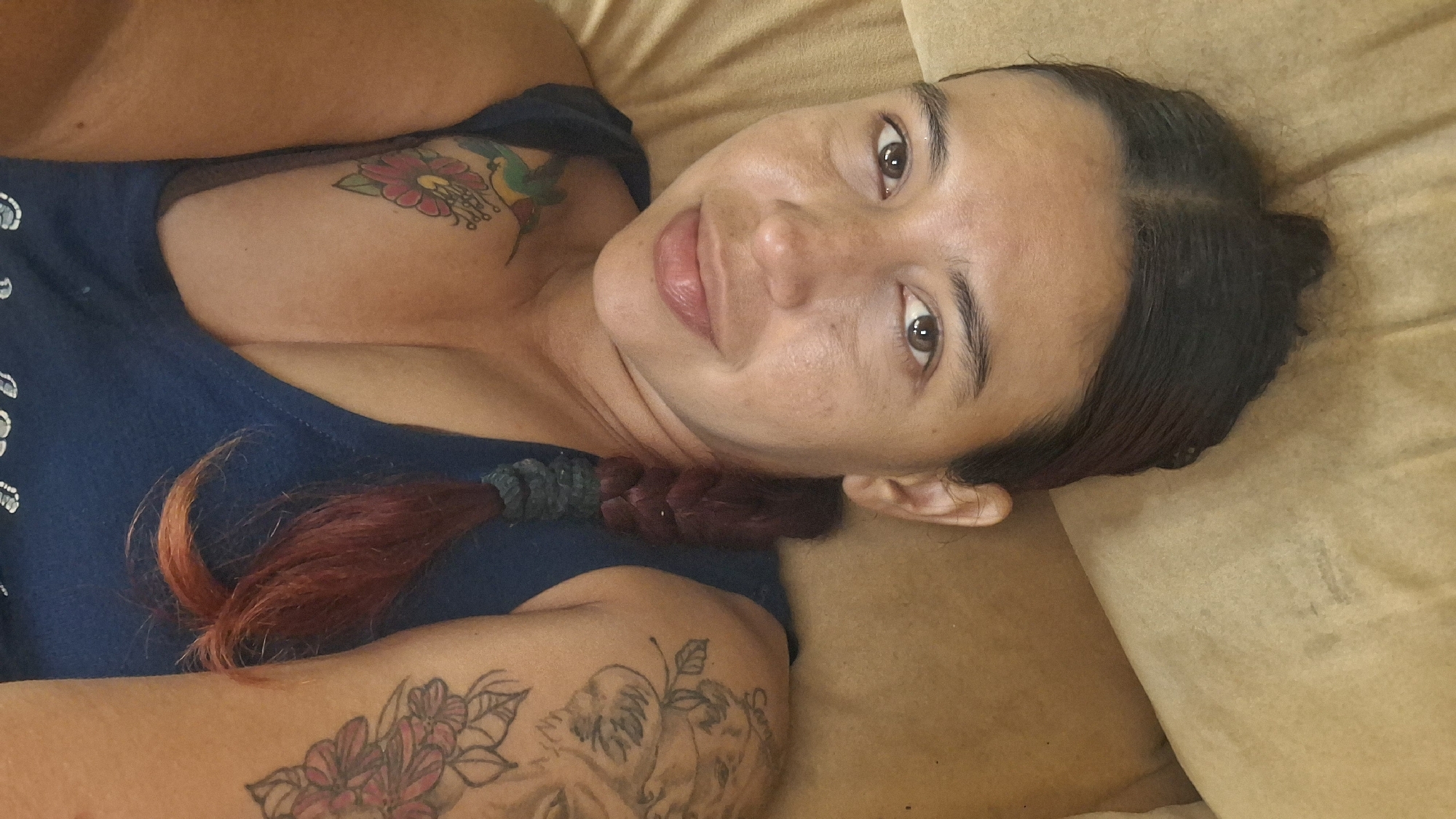 A medium shot captures a woman with medium-toned skin and dark hair styled in a bun and a braid. She is lying down, with her head resting on a textured tan surface. Her eyes are open, and she is looking at the camera with a slight smile. She is wearing a dark blue tank top, and has tattoos on her left chest and right arm. The tattoos on her chest depict a hummingbird and a flower, while the one on her arm features flowers. The setting appears to be indoors, with the tan surface possibly being a couch or bed. There is no indication of the specific location in Vallecito, Venezuela.