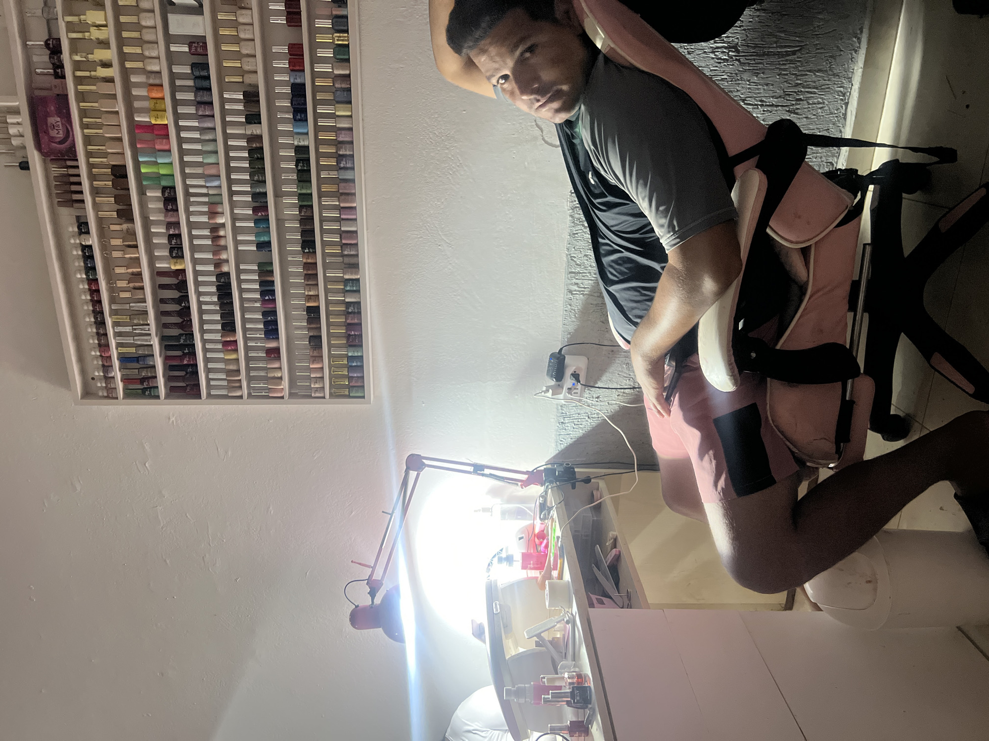 In Juan Griego, Venezuela, a man is reclining in a pink, office-style chair in what appears to be a nail salon. The chair is angled away from the camera, and the man is looking towards it with his head resting on his arm. He is wearing a grey t-shirt and pink shorts. A shelf filled with various colored nail polishes is mounted on the wall behind him. In the foreground, a pink desk is visible, equipped with a bright red adjustable lamp, a UV nail lamp, and an assortment of nail care products. A white trash can is placed next to the chair. The lighting is a mix of bright artificial light from the lamp and ambient room light.