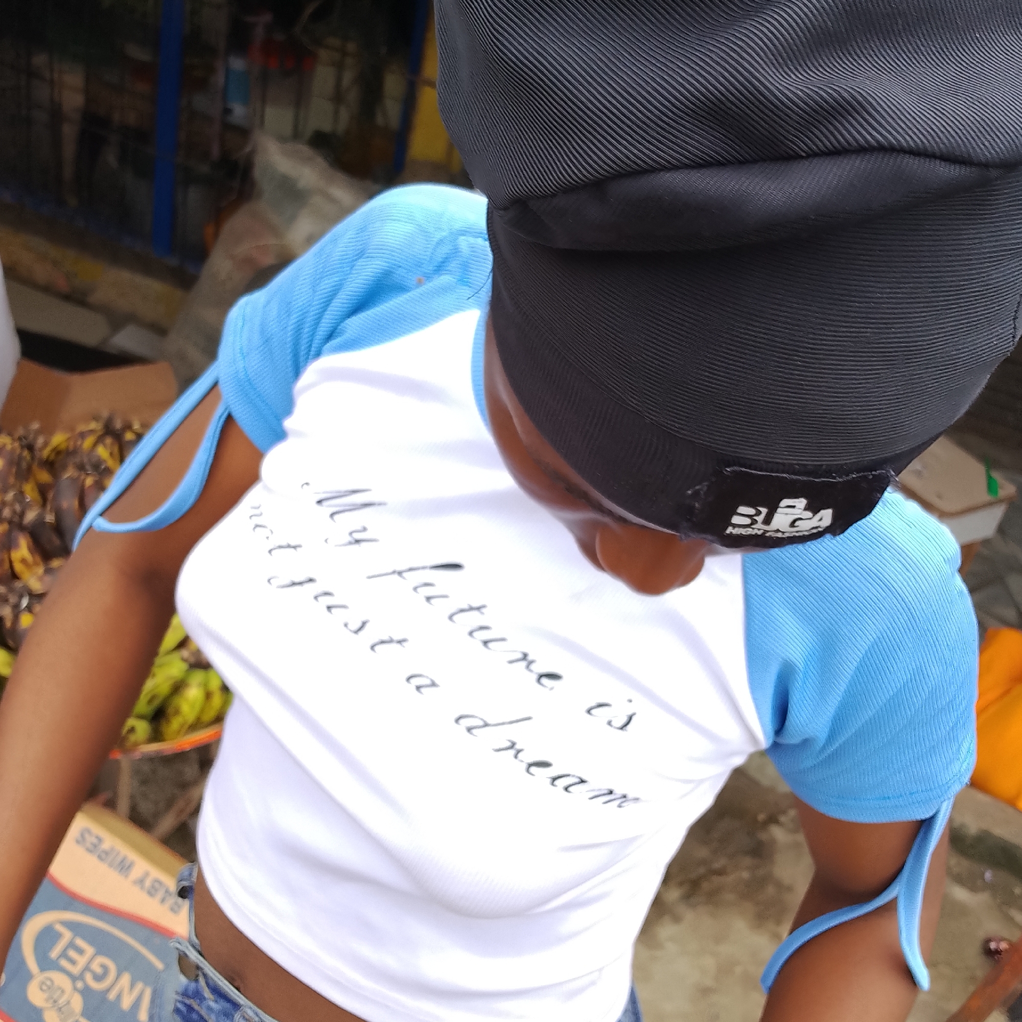 In Ado, Nigeria, a person is shown from a high angle perspective, wearing a black du-rag and a white t-shirt with blue sleeves. The t-shirt has cursive text printed on it that reads "My future is not just a dream." A black tag with white lettering, possibly a brand logo, is attached to the sleeve near the neckline. The person is standing in what appears to be an outdoor market setting, with a pile of bananas visible in the background, and a cardboard box with "BABY WIPES" printed on it in the lower left corner. The ground is concrete. The lighting suggests it is daytime.