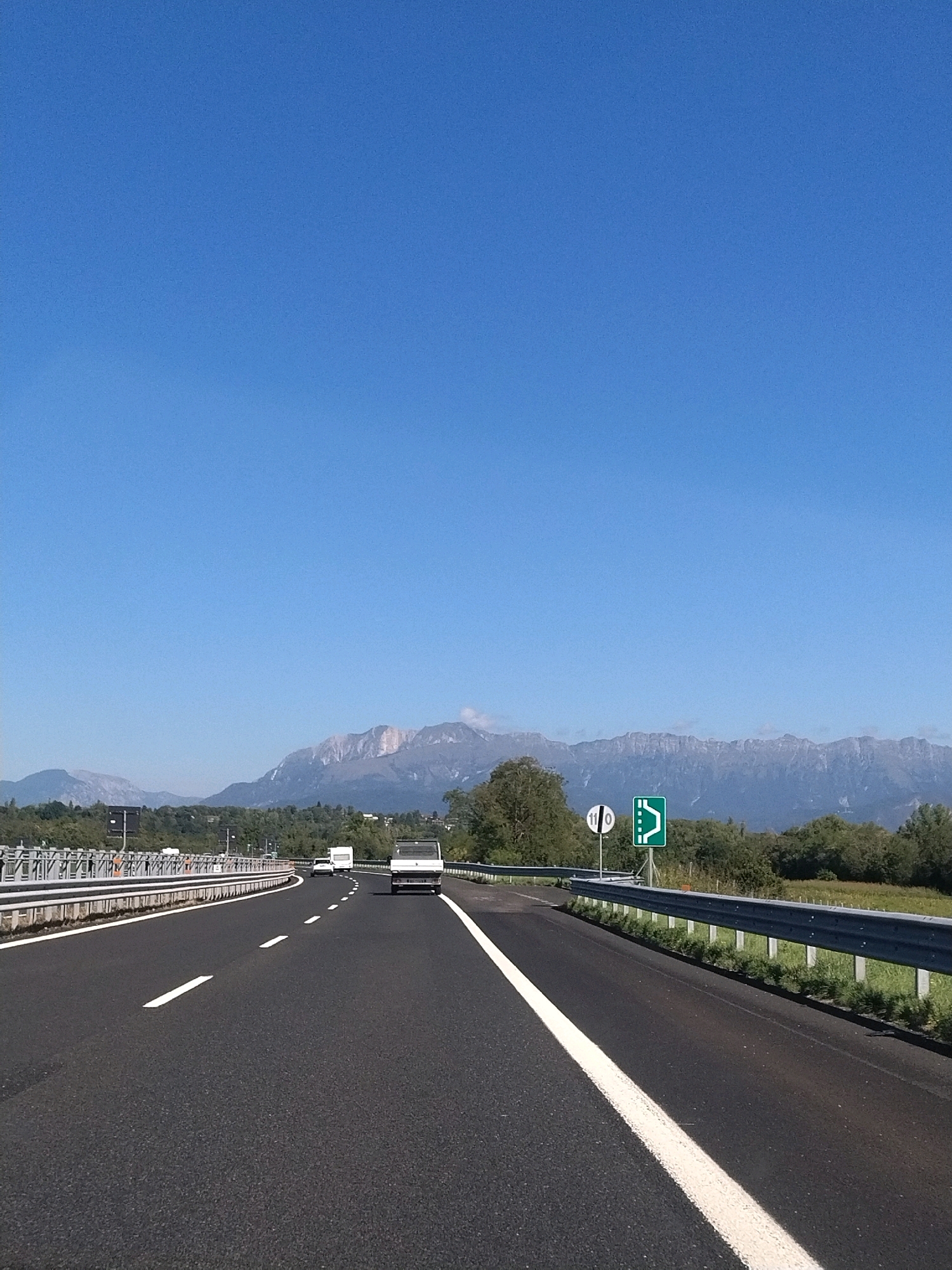 The image shows a highway scene in the city of Castellerio, Italy, with a clear blue sky overhead and mountains in the background. The highway is asphalt with white lane markings. There are several vehicles on the road, including a white camper van, a white truck, and a white car. There are also guardrails along the side of the road and some signage. The landscape is characterized by trees and vegetation. The overall impression is one of a sunny day on an Italian highway.