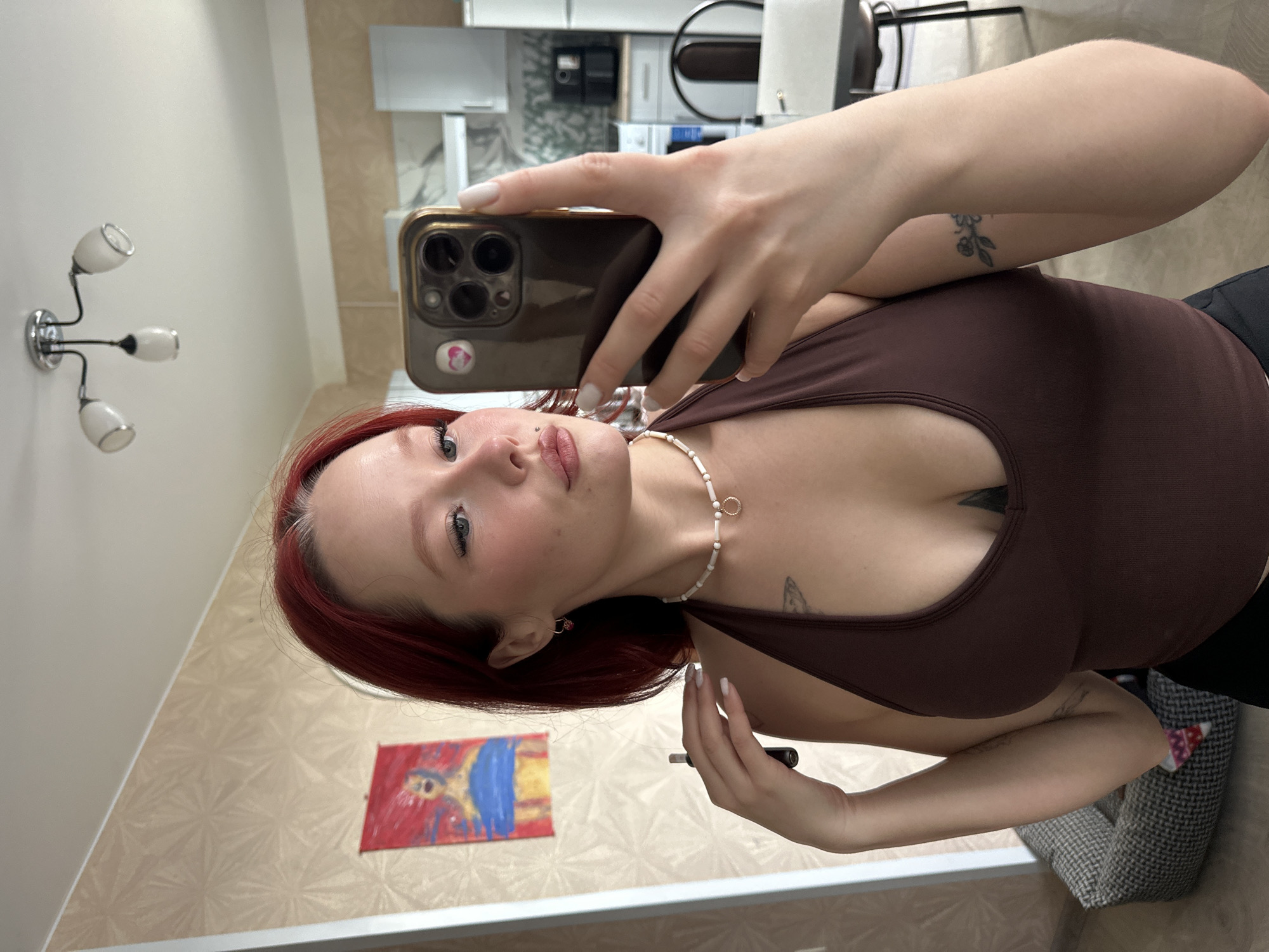 A young woman with red hair and a nose ring takes a selfie in what appears to be a residential kitchen. She is wearing a brown tank top and a beaded necklace. In her right hand, she holds a smartphone with a gold case, which she uses to capture the reflection. A painting hangs on the wall behind her, and a light fixture is visible on the ceiling. The background also shows kitchen cabinets, appliances, and furniture, including a chair and table.
