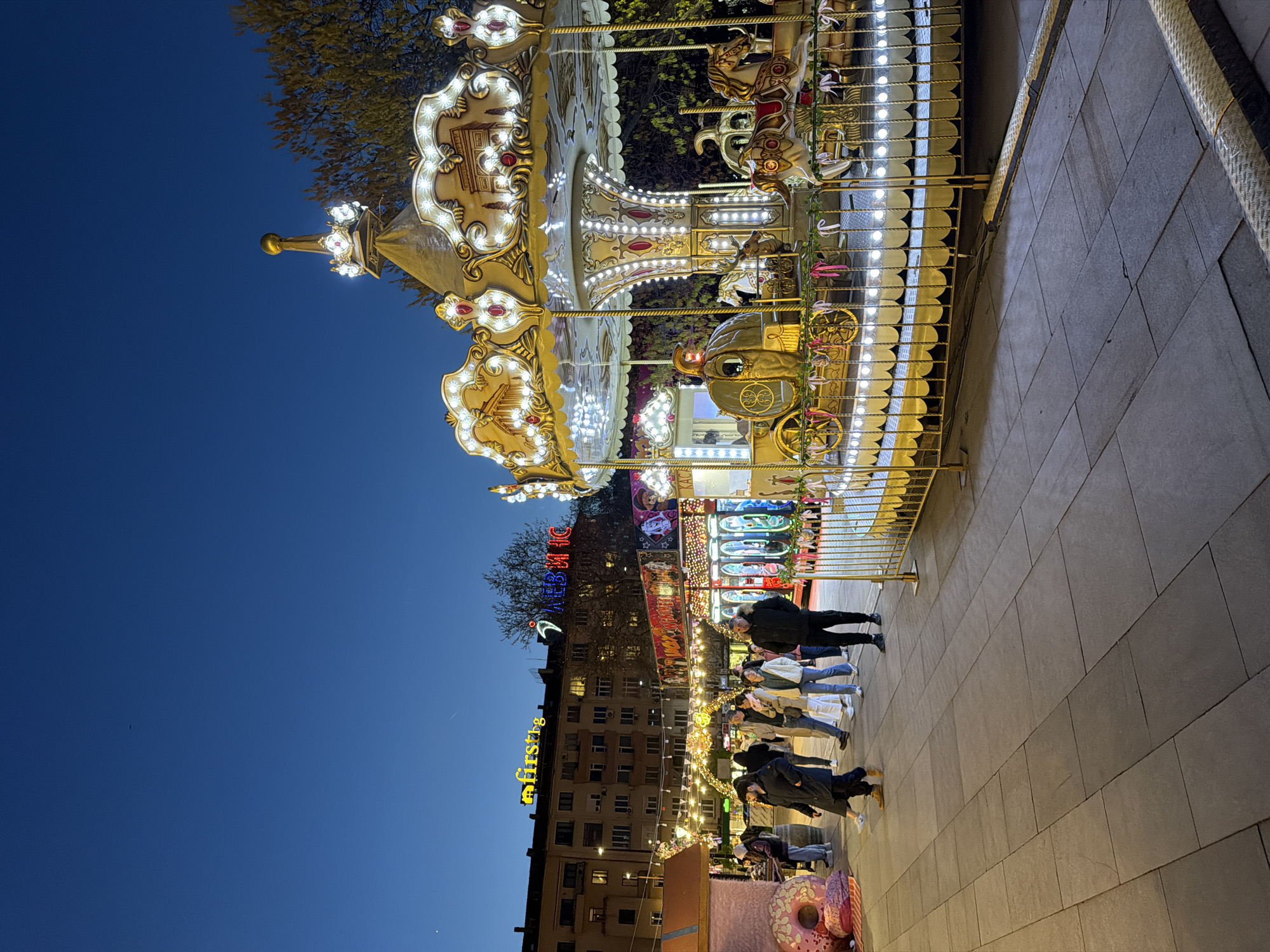 The image captures a vibrant nighttime scene, likely at a public fair or market, set against the deep blue of a twilight sky. In the foreground, a grand, ornate carousel dominates, brightly illuminated with numerous warm white and golden lights that highlight its intricate decorative details, including horses and a carriage. A golden railing separates the carousel from a paved pedestrian area.
Several casually dressed people are visible on the paved ground, standing or strolling, observing the attractions. Their postures suggest a relaxed and leisurely atmosphere.
Beyond the carousel, the background features more brightly lit stalls or rides, adding to the festive ambiance. Further back, multi-story buildings line the street, with some windows aglow, suggesting urban surroundings. Sparse trees are visible between the buildings and the fairground attractions. The ground is paved with large, dark grey tiles.
Notable details include a large yellow sign on one of the buildings spelling "first8". Above it, a neon sign in Cyrillic letters reads "ТАЛКЕВИС" (TALKEVIS), with the last letter partially cut off. Another partial neon sign above this one appears to show "COCA КОЛА" (Coca Cola) in Cyrillic, providing a location cue consistent with Sofia, Bulgaria. The ample artificial lighting contrasts with the deep blue sky, indicating either dusk or early night. The overall impression is one of festive entertainment and public enjoyment.