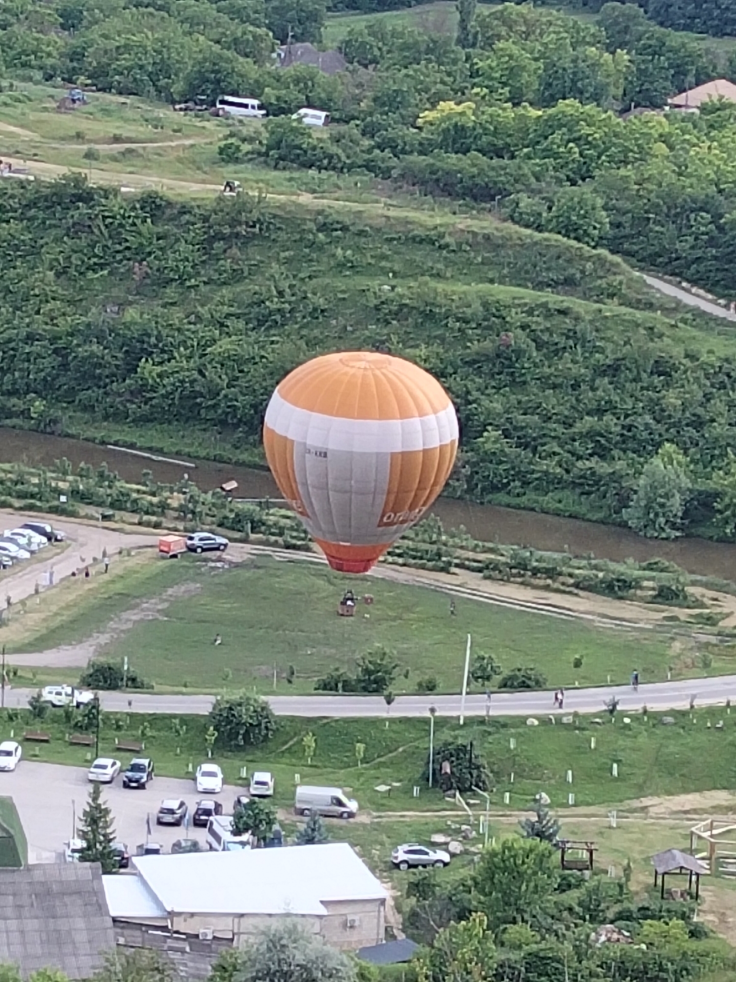 A hot air balloon, with orange and white panels, drifts over a verdant landscape. The balloon is the central subject, appearing to be either ascending or descending, with a basket visible beneath it. The scene is a rural setting in Butuceni, Moldova, characterized by rolling green hills densely covered with trees. A narrow, brown river or stream snakes through the middle ground.
In the foreground and midground, there are signs of human activity and infrastructure. A parking area is visible, filled with various vehicles including cars, SUVs, and a van. A white building with a light-colored roof is present in the lower left. Roads and pathways wind through the grassy areas, with a few benches placed along a path. Several people are scattered throughout the scene, some walking along the road, others in the grassy areas. There is also a small wooden structure with a roof, possibly a picnic shelter.
The overall impression is of a tranquil, daytime scene, likely during pleasant weather, conducive to outdoor activities like hot air ballooning. The lush greenery suggests a spring or summer season. The image lacks any discernible text that can be read clearly.