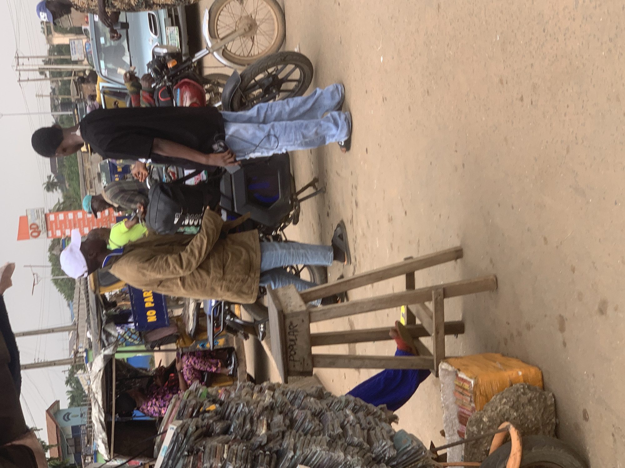 The image captures a street scene in Shagamu, Nigeria, likely a marketplace given the presence of vendors and assorted goods. In the foreground, a wooden stool with the word "POPULAR" inscribed on it stands on the dusty, light brown road. To the left, a large stack of what appears to be packaged goods, possibly books or DVDs, is piled high. A person's leg, wearing a red sandal, is visible beneath the stool.
In the middle ground, several individuals are present. A man wearing a brown jacket and a white cap stands near the stool, facing right. Behind him, a woman in a vibrant purple and pink patterned outfit sits inside a covered vehicle, possibly a tricycle or a small bus. Further back, another man in a black t-shirt and light blue jeans stands facing left, holding a device that may be a mobile phone. A red and black motorcycle is parked to his right.
In the background, a light blue car is visible, and behind that, more vehicles and buildings. A blue sign with yellow lettering that reads "NO PAR" is partially visible. The environment suggests a busy urban setting with a mix of commerce and traffic. The overall lighting indicates it is daytime, with overcast or diffused sunlight.