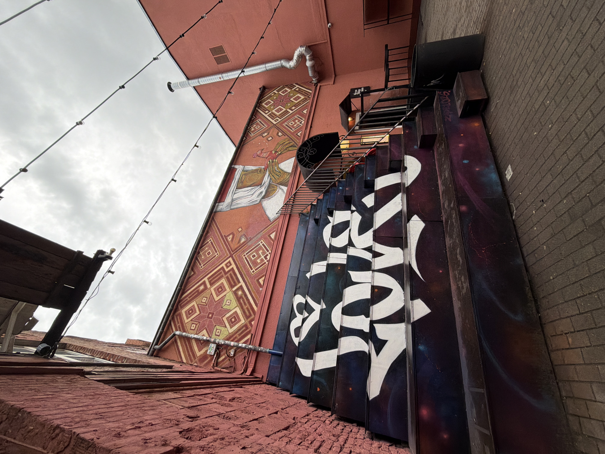 A low-angle photograph captures an alleyway between brick buildings, with a view extending upwards towards the sky. The buildings' facades are a mix of textures and colors. A coral-colored building features a patterned mural of figures in robes, above which there's a vent pipe and a string of lights against a cloudy sky. Adjacent to the mural, stairs lead up to an unseen higher level, next to a wall with a dark, space-themed mural with white graffiti lettering on it. The alley floor is brick-paved.