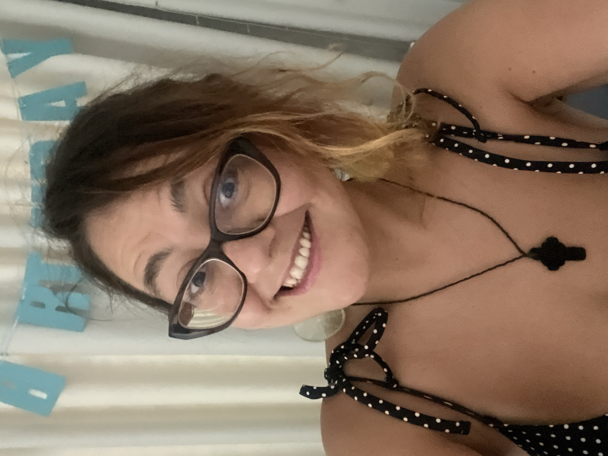 The image is a medium shot selfie of a woman with brown hair and glasses, smiling at the camera. She is wearing a black and white polka dot top and a black pendant necklace. Behind her, a white curtain is visible, and a blue banner with "HAPPY BIRTHDAY" written on it in cut-out letters is hanging on the curtain. The lighting suggests it is daytime, and the background is slightly blurred, indicating a shallow depth of field. The woman appears to be in a domestic setting, possibly a home or a hotel room. There is no discernible information in the image that indicates a specific location in Nha Trang Ward, Vietnam.