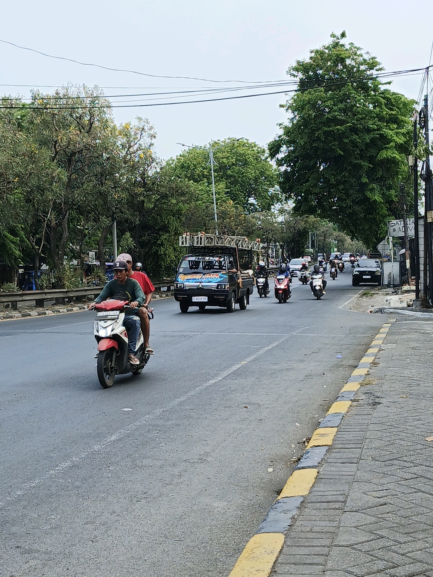 The image depicts a street scene in Ngaban, Indonesia. A two-lane asphalt road runs horizontally across the frame. On the left side of the road, there is a sidewalk with a curb marked by yellow and gray blocks, bordered by trees and a metal barrier. On the right side of the road, there is a wider sidewalk made of interlocking concrete tiles, also bordered by trees and a fence.
Several vehicles are present on the road. In the foreground, a white and red motorcycle is ridden by two men. The driver wears a green long-sleeved shirt, blue jeans, and a dark baseball cap, while the passenger wears a red shirt and a pink cap. Further down the road, a small blue truck with a ladder rack is visible, carrying construction materials. A procession of motorcycles is following behind the truck, with riders wearing helmets and casual clothing. A black car is also seen in the distance on the right lane.
The environment appears to be urban or suburban, with mature trees lining both sides of the road. Overhead power lines are visible against a hazy, overcast sky. A sign with text, partially obscured, is mounted on a pole on the right side of the road, indicating a commercial establishment. The overall impression is of a busy street on a moderately cloudy day.