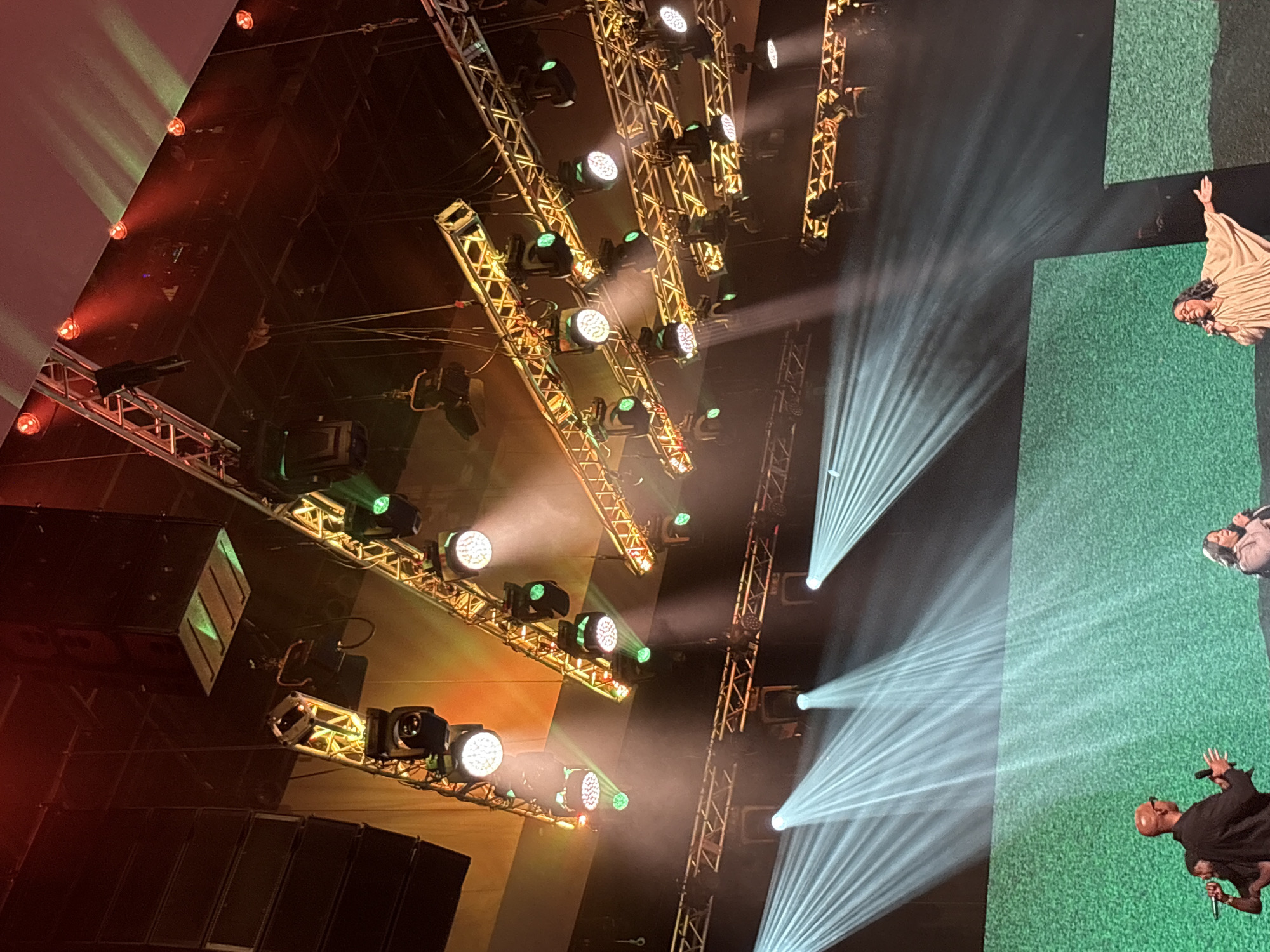 The image depicts a stage setup, likely in Dallas, Texas, USA, with three individuals visible on stage. The backdrop is a textured green screen or wall. In the foreground, a man with a shaved head and glasses is speaking into a microphone, holding it with his right hand while gesturing with his left. To his left, another person is partially visible, also with a microphone. To the right, a woman with long dark hair and glasses is singing into a microphone, wearing a light-colored, flowing garment.
Above the stage, a complex lighting rig is suspended from trusses. Numerous spotlights and stage lights are visible, some emitting bright white beams that create dramatic rays in the smoky atmosphere, while others cast warm orange and red glows. Large black speaker arrays are also mounted above the stage. The overall impression is of a live performance or event in progress.