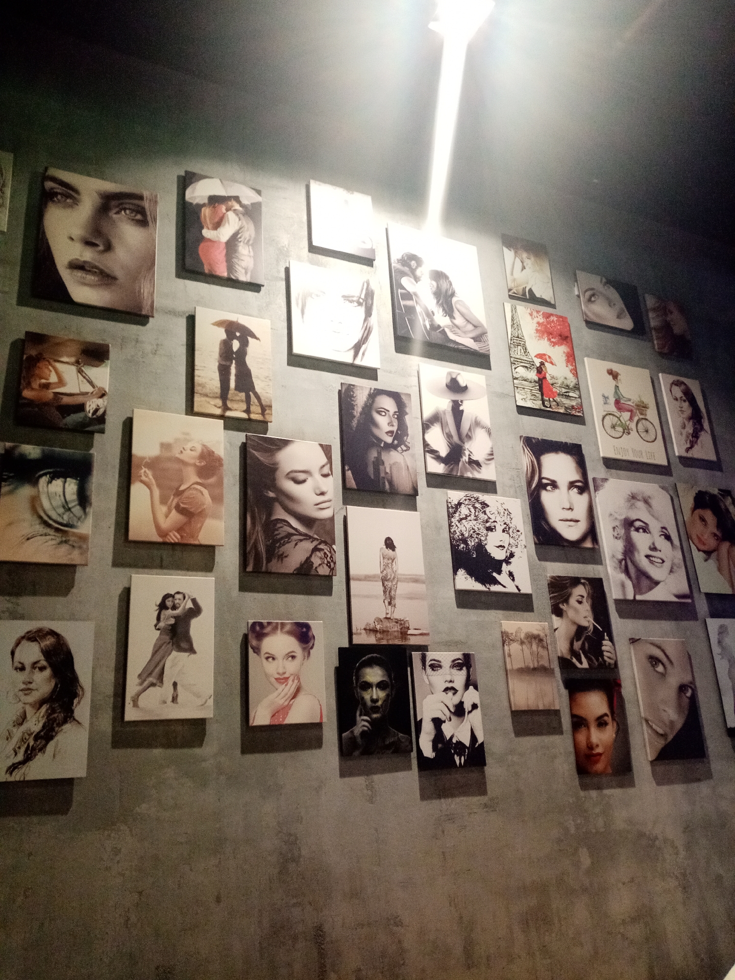 The image shows a wall in what appears to be an interior space, possibly a restaurant or bar. The wall is textured, resembling concrete or stucco. Displayed on the wall is an assortment of approximately 30 framed pictures of varying sizes, mostly depicting portraits of women and romantic scenes. Many are in black and white, while some feature color. The light source is above and casts a bright glow, partially obscuring the top portion of the wall. The scene is dimly lit overall, creating a slightly somber or artistic atmosphere. The location is Izhevsk, Russia.