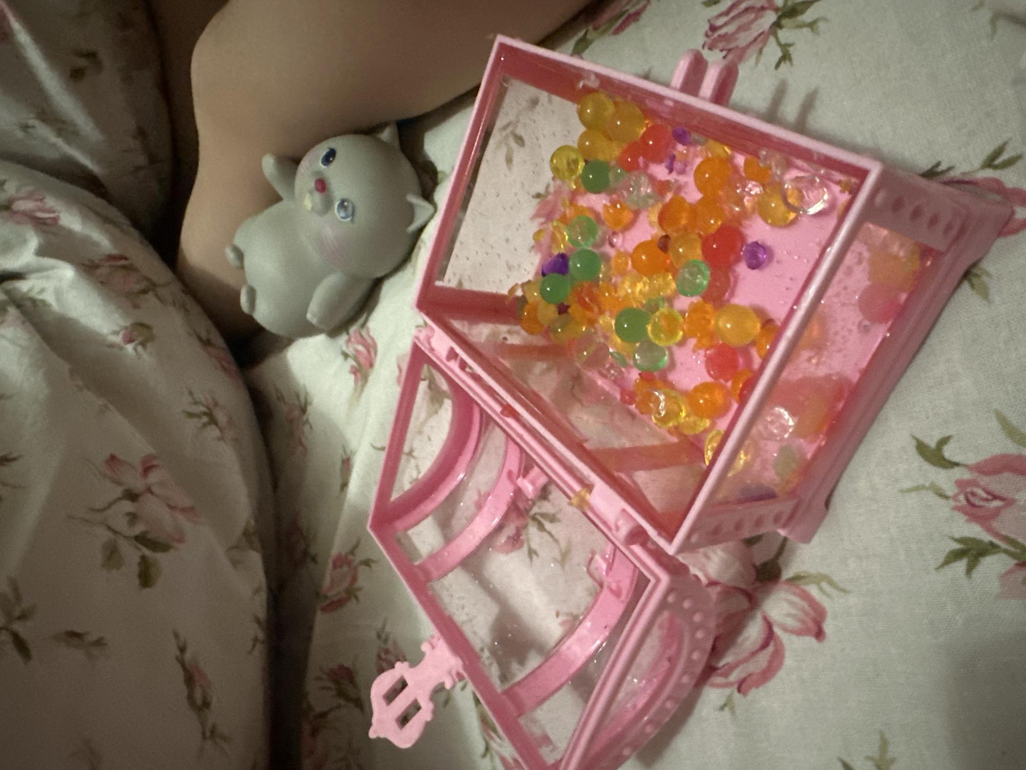 This image appears to be taken indoors, likely on a bed. The floral patterned bedding is predominantly white with pink and green floral designs. A pale-skinned person's leg and arm are present on the left side of the frame. Next to the leg is a small gray toy figurine, resembling a stylized animal with large eyes. To the right, a pink toy treasure chest is open. Inside the chest, there are a number of colorful water beads. The background is blurred, giving the image a shallow depth of field. The location is not discernable from the image.