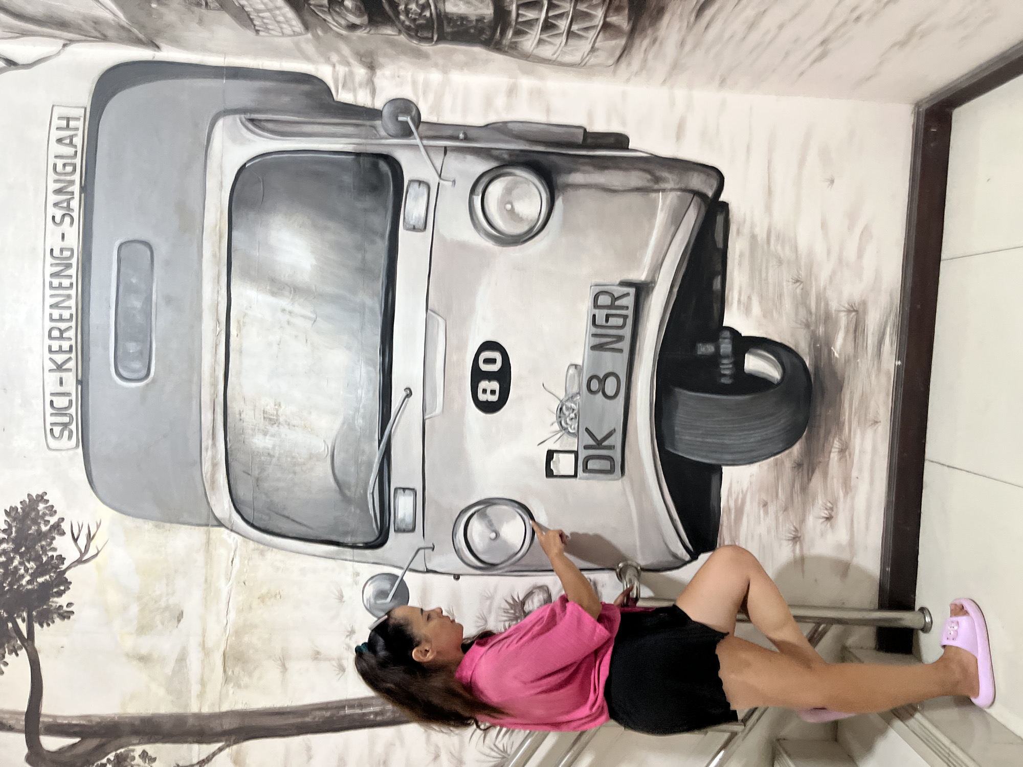 The image shows a woman posing in front of a large mural of a vintage vehicle on a wall. The mural is in grayscale and depicts the front of what appears to be a three-wheeled car, possibly a tuk-tuk. The vehicle has a license plate that reads "DK 8 NGR". There is also text on the side of the vehicle that says "SUCI-KERENGNG-SANGALAH".
The woman is wearing a pink t-shirt and black shorts, with pink slippers on her feet. She is leaning on a metal railing, with her right arm extended, pointing towards the mural. She appears to be interacting with the artwork, creating a playful or engaged pose. The mural also features elements of nature, such as trees and branches, painted in a similar grayscale style.
The setting appears to be an indoor space, possibly a hallway or a common area, with a tiled floor and a doorway visible on the right side of the image. The lighting suggests it could be daytime. The overall impression is that of a tourist or local enjoying a piece of street art in Kuta Selatan, Indonesia. The woman's expression is neutral and focused on her pose.