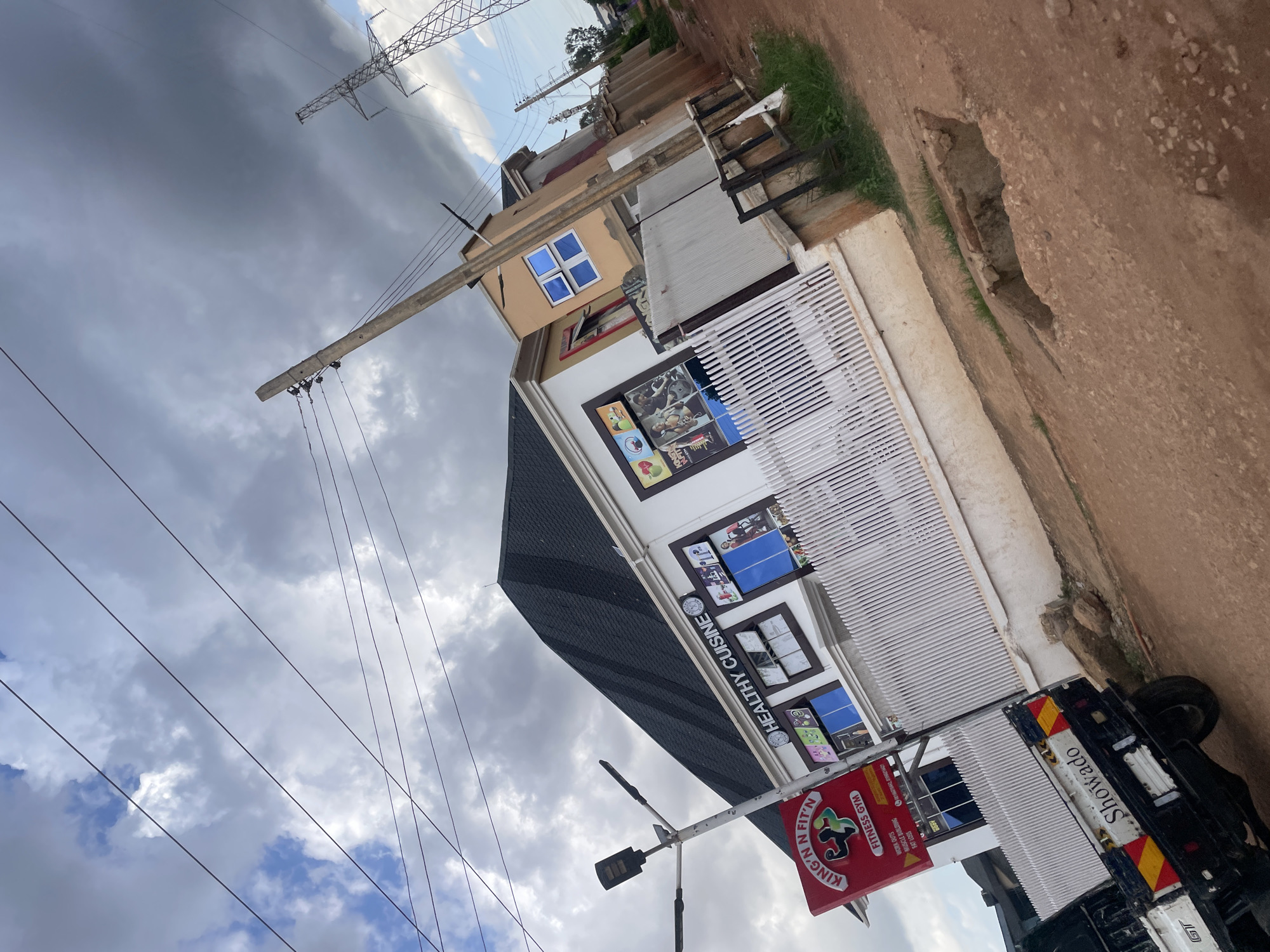 The image shows a street scene in Gura Top, Nigeria, under a cloudy sky. A white building with a dark, sloped roof is prominently featured, housing a business labeled "HEALTHY CUISINE" above several windows. To the left of this building, a red sign for "KING 'N' FIT'N FITNESS GYM" is visible, with graphics of a muscular figure. A street light stands near the gym sign. In the foreground, the rear of a white and black pickup truck is visible, with "Showado" written on its tailgate. The ground is unpaved, with loose dirt and rocks, and a hole in the road is present. Electrical poles and wires crisscross the sky, and a large transmission tower is seen in the distance.