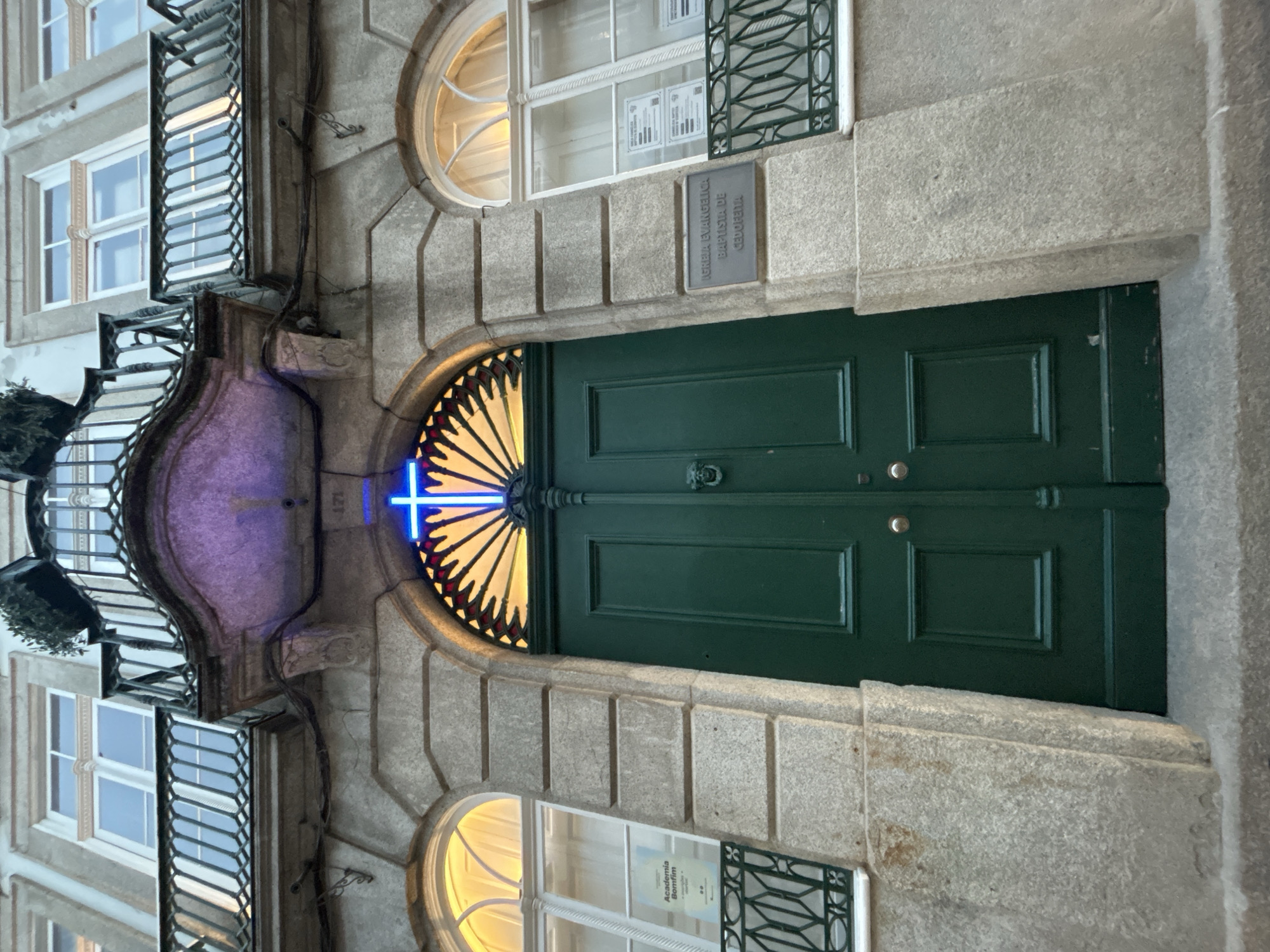 This is a low-angle, medium shot of the exterior of a stone building with a large, dark green double door in the center. Above the door is an arched window with a fan-shaped stained-glass design and a bright blue neon cross illuminated in the center of the arch. To the left and right of the door are arched windows with white frames and yellow interior lighting. The building has ornate balconies with black metal railings on the upper floors. A sign on the right side of the building reads "IGREJA EVANGÉLICA BAPTISTA DE CEDOFEITA". The stone facade of the building is rough and textured.