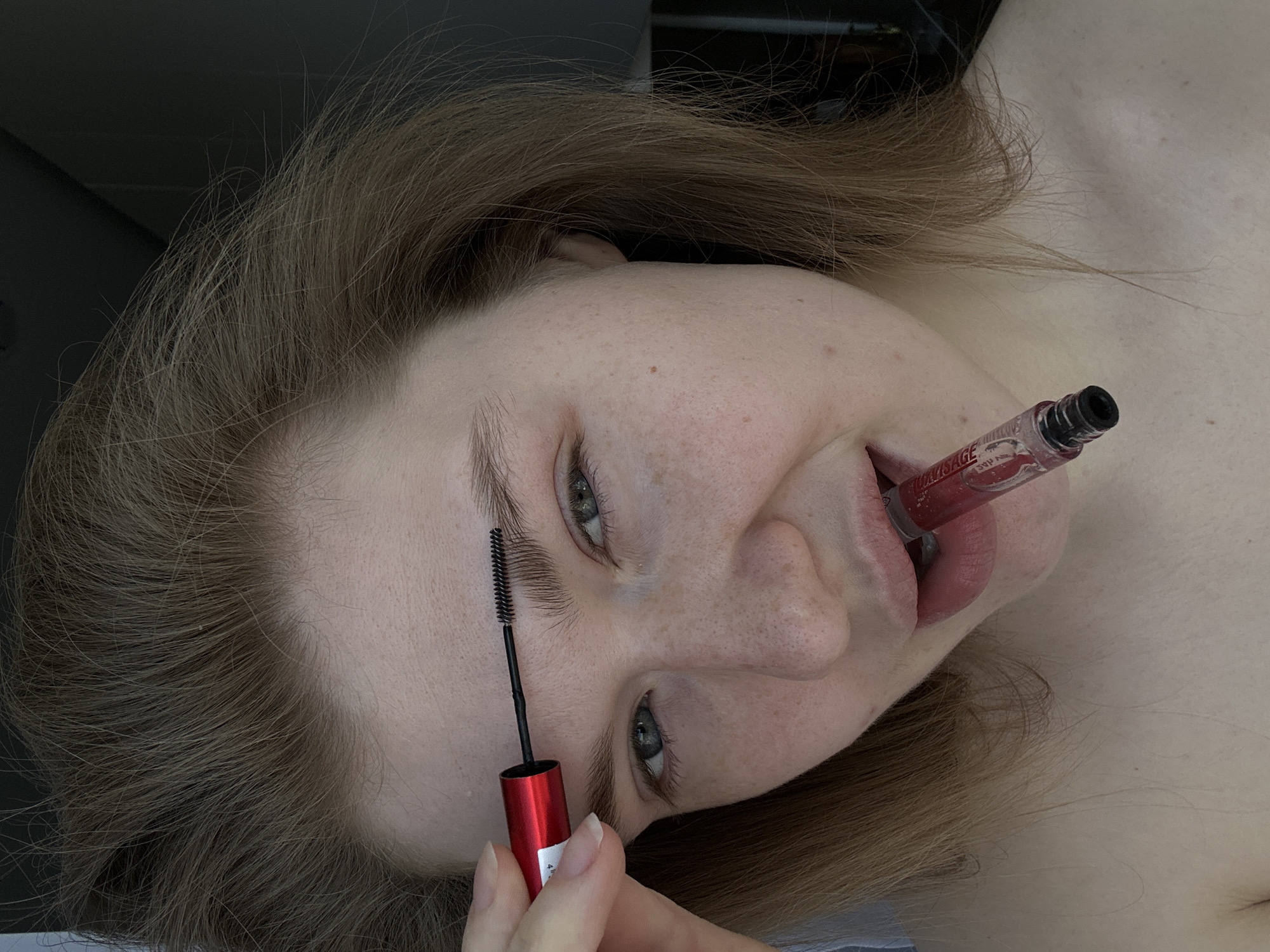 The image presents a close-up, eye-level portrait of a young adult female captured indoors in Kazan, Russia. Her fair skin displays scattered freckles across her nose and cheeks. She possesses light-colored eyes and light brown hair, which appears to be unstyled and cascades around her face. She looks directly towards the viewer with a subtle, possibly playful, expression.
Her right hand holds a red and white cosmetic applicator featuring a black bristled brush, positioned above her left eyebrow as if in the process of grooming or applying product. Concurrently, a clear tube of red liquid cosmetic, appearing to be lip gloss or liquid lipstick, with the partially visible word "MASSAGE" on its label, is held horizontally between her slightly parted lips. Her bare shoulders are visible in the lower portion of the frame, suggesting a lack of upper body clothing. The background is dark and out of focus, indicating an undifferentiated indoor environment.