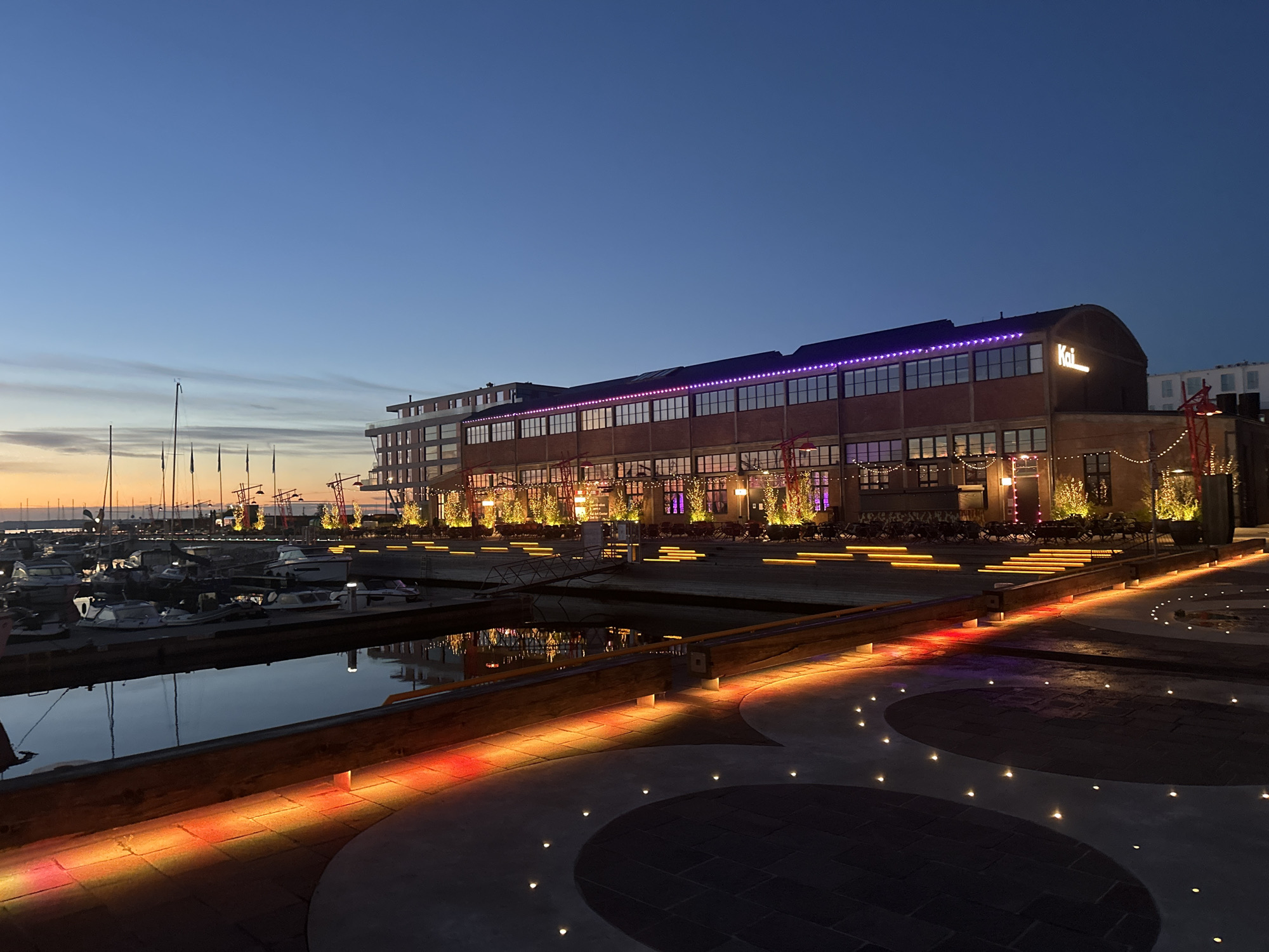 This image depicts the waterfront area in Tallinn, Estonia, during twilight. A modern brick building with large windows, illuminated by purple and yellow lights, dominates the background. The building features a prominent "Kai" sign. In the foreground, a marina with several docked boats is visible, their reflections shimmering in the calm water. The paved walkway in front of the building is lined with colorful lights, casting a vibrant glow on the ground. The sky transitions from a pale orange at the horizon to a deep blue overhead. No people are present in the scene.