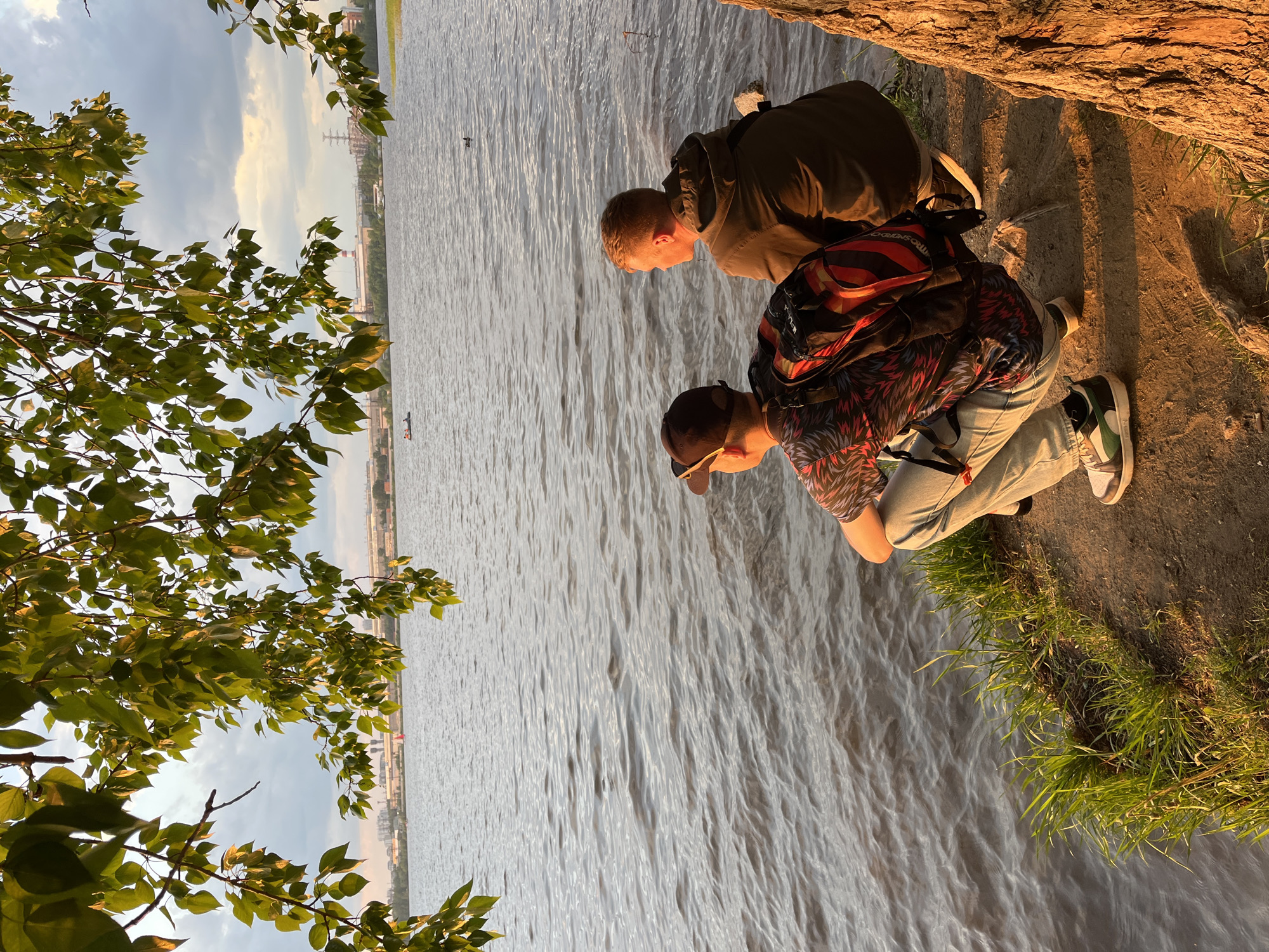 Two people are near a body of water. The person closest to the viewer wears a red, black, and white patterned shirt, light-colored jeans, and green and white sneakers. They have a backpack, and a dark-colored cap. The second person wears a dark green jacket. The water is gray and has ripples. In the distance across the water is what appears to be the city of Yekaterinburg, Russia, identified by the presence of industrial structures. In the foreground, there is some sparse greenery and a dirt embankment. The scene is lit by a warm light, suggesting either sunrise or sunset.