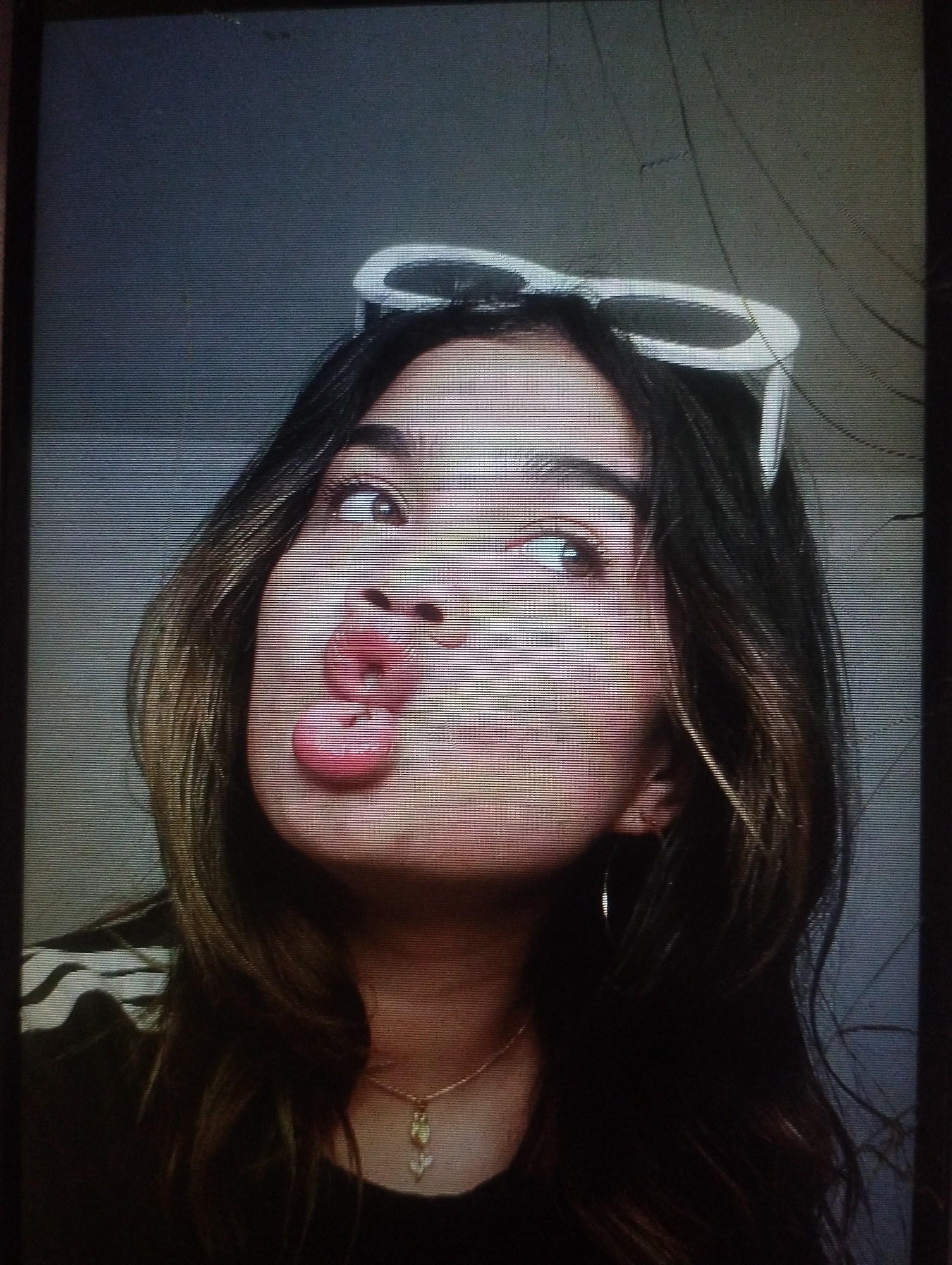 A young woman with long, dark hair poses for a photo. She has white sunglasses perched on her head. She's making a "duck face." She's wearing a dark shirt and a delicate necklace. The background appears to be a plain wall. No identifiable details indicate the location is Punta de Piedras, Venezuela.