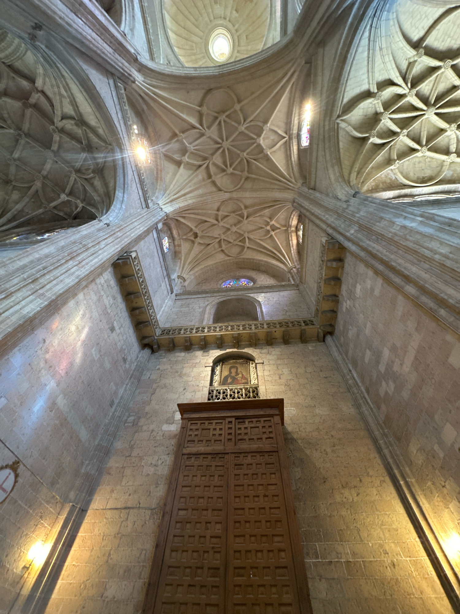 The image depicts the interior of a cathedral in the city of La Lastrilla, Spain. The view is from a low angle, looking upwards towards a highly ornate vaulted ceiling. The ceiling is constructed of stone, with intricate geometric ribbing and circular patterns. Sunlight streams through high arched windows, casting colored light onto the stone walls.
In the center of the lower portion of the image, a large, dark wooden cabinet with a highly patterned door is visible, reaching from the floor upwards towards the vaulted ceiling. Above this cabinet, a framed religious artwork is mounted on the wall, depicting a figure, likely Mary, holding a child. This artwork is set within a decorative metal railing. The walls are constructed of large, rough-hewn stone blocks. Lights are visible on the walls, casting illumination. The overall impression is one of grandeur and historical significance.