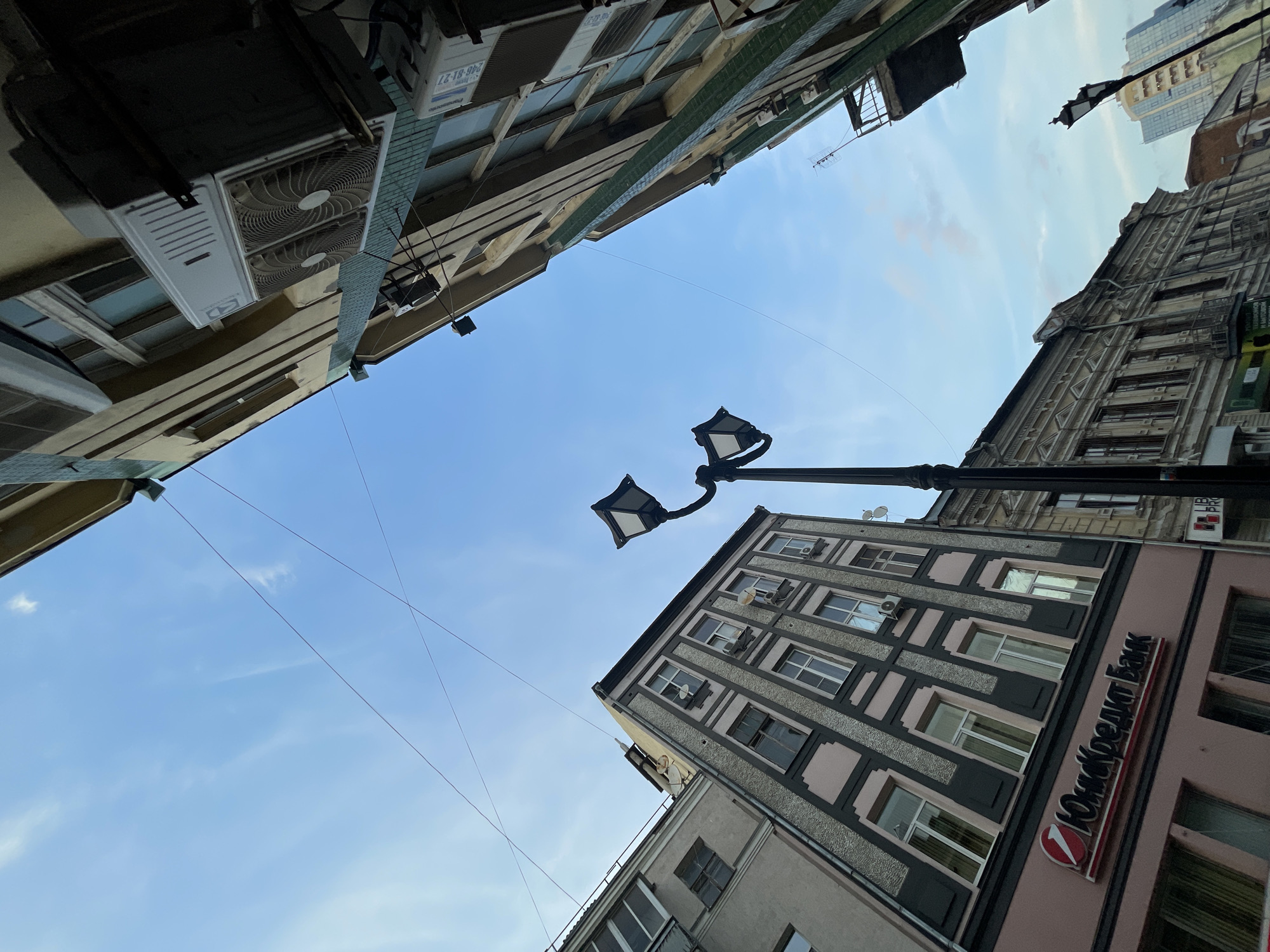 The image shows a street scene in Rostov-on-Don, Russia, with a low-angle perspective looking up at buildings and the sky. The buildings are a mix of architectural styles, with older, ornate facades and more modern structures. One building prominently features the logo for "Юникредит Банк" (UniCredit Bank). Several air conditioning units are visible on the exterior of a building. Overhead, power lines crisscross the sky, and a street lamp is visible in the foreground. The sky is blue with some wispy clouds. There are no people visible in the image.