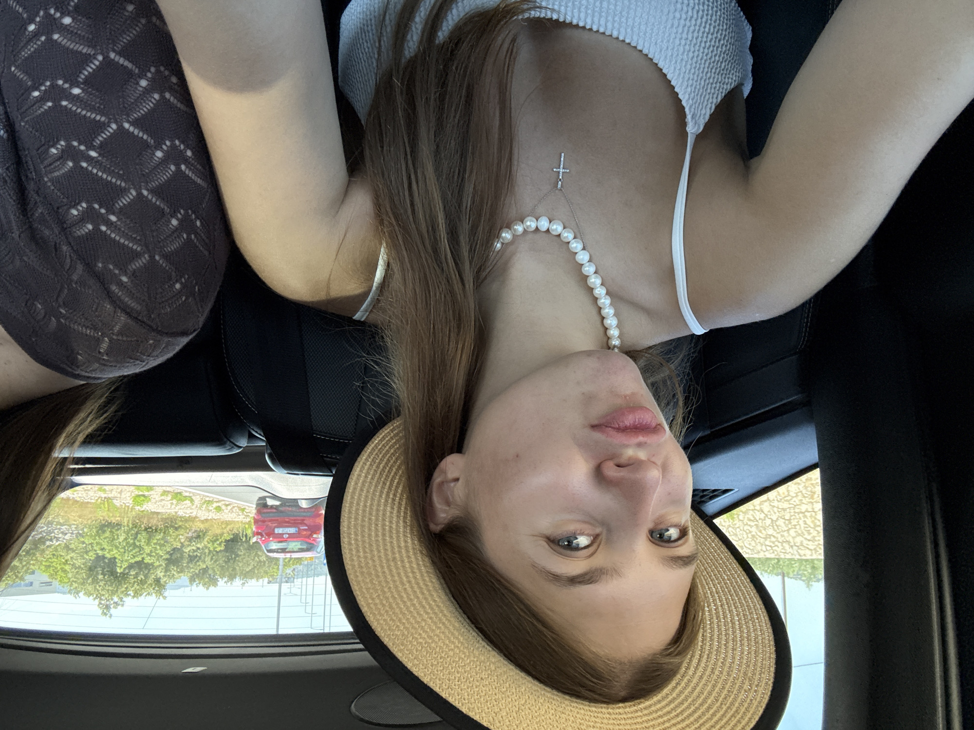 A young woman with long, light brown hair, wearing a straw hat, a white textured top, and a pearl necklace with a silver cross pendant, takes a selfie in the back seat of a car. She is pouting and looking towards the camera. Her left arm is visible, resting against the car's interior. Another person's shoulder and arm, wearing a dark, patterned top, are visible on the right side of the frame. Through the car windows, a bright, sunny day is visible, with a red car driving on a road in the distance, surrounded by green trees and vegetation. The scene suggests a casual road trip or outing, possibly in or near Grad Šibenik, Croatia, given the Mediterranean-like scenery.