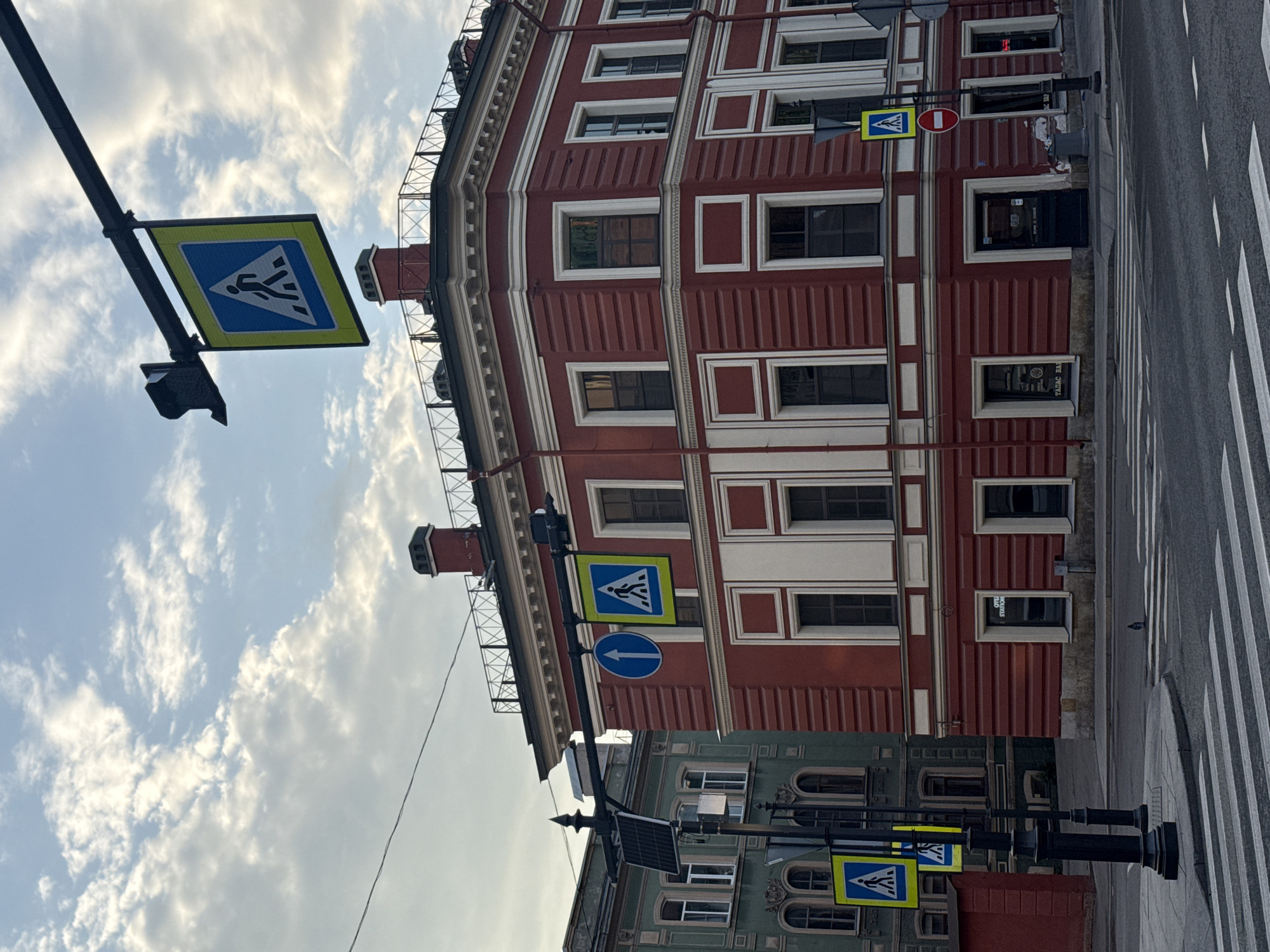 The image depicts a street scene in Saint Petersburg, Russia, featuring a red building with white trim and a crosswalk. Street signs indicating a pedestrian crossing are visible on both sides of the street. The sky is cloudy.