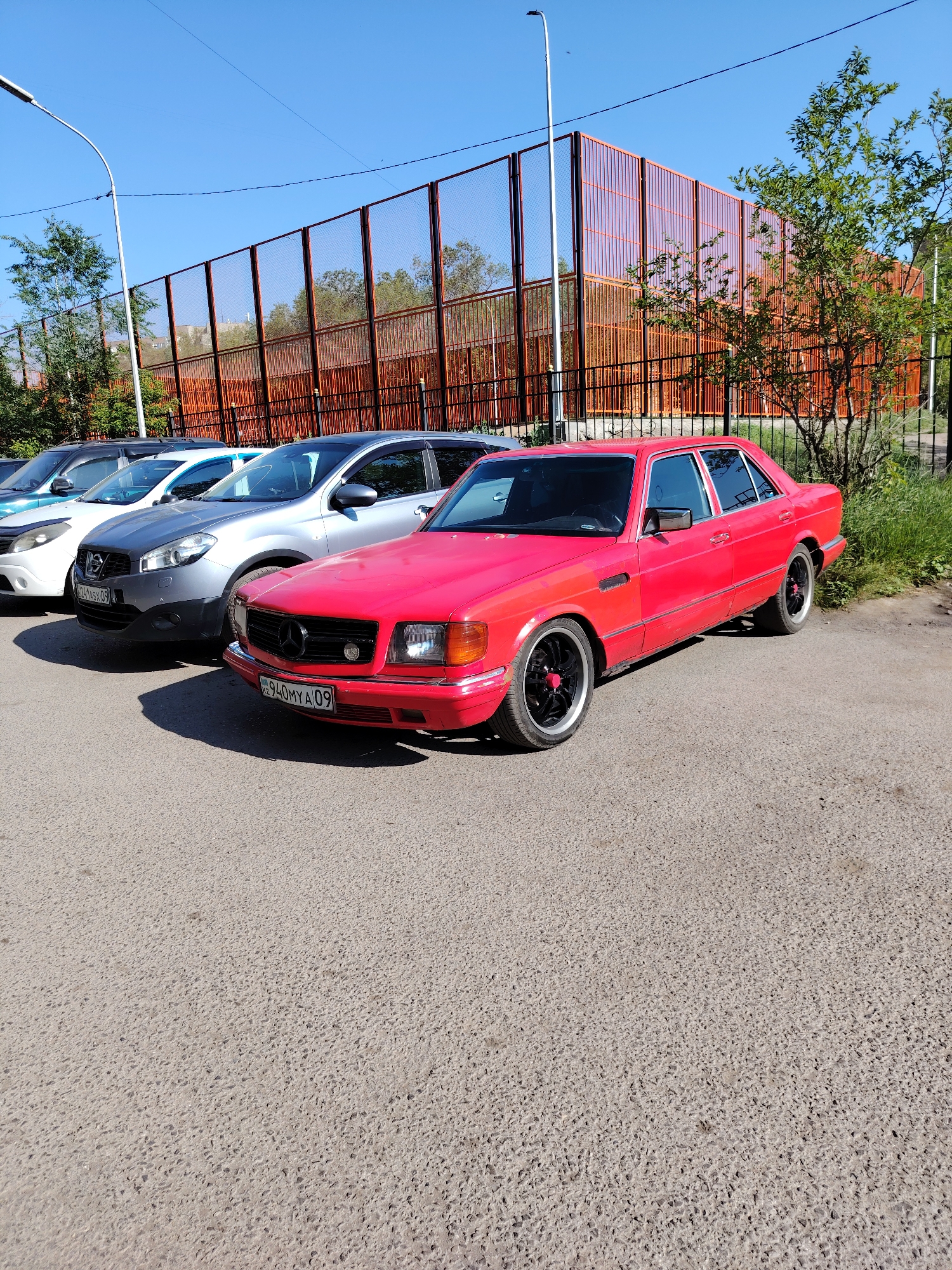 A bright red Mercedes-Benz sedan is parked on an asphalt surface. The car has black rims with red accents. In the background, there is a fence with orange panels and a chain-link section. Trees and grass are visible behind the fence. Several other cars are parked in a row to the left of the red Mercedes. The sky is clear and blue. The license plate on the red Mercedes-Benz reads "KZ 940MYA 09". The image appears to have been taken on a sunny day. The overall scene depicts a parking lot area, likely in an urban or suburban setting.