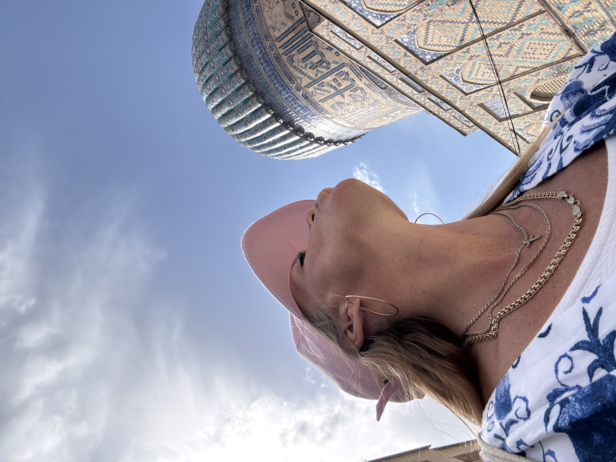 A low-angle shot captures a woman wearing a pink hat and a white and blue floral patterned shirt. She is gazing upwards at a large, ornately decorated structure featuring intricate blue and tan mosaic tiles, presumably a building of cultural or historical significance. The sky is visible in the background, showing a mix of clear blue and cloudy areas. The woman is also wearing multiple gold necklaces and earrings. Based on the architectural style, this image was likely taken at Registan Square in Samarkand City, Uzbekistan.