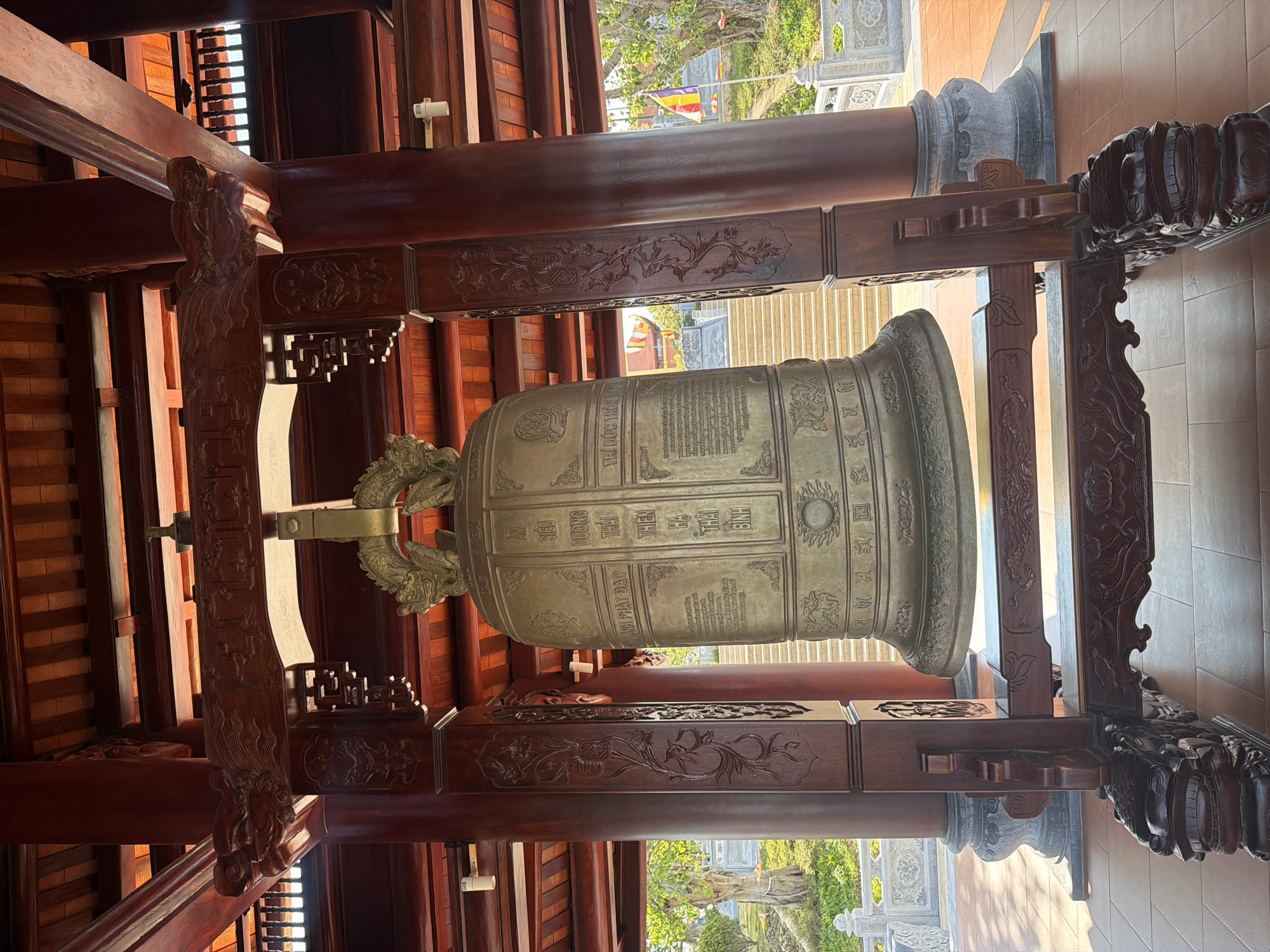 A large bronze bell with inscriptions hangs suspended within an ornate, traditional wooden structure. The bell is positioned centrally in the frame, showcasing its detailed surface, including the decorative elements and the panels of text. The wooden structure is constructed from dark, polished wood, featuring intricate carvings and designs that suggest a cultural or religious significance. The environment is open, with glimpses of exterior elements visible beyond the structure, including steps, foliage, and distant flags. The overall setting appears to be outdoors, possibly within a temple or public space. The image was taken in Nam Nha Trang Ward, Vietnam.