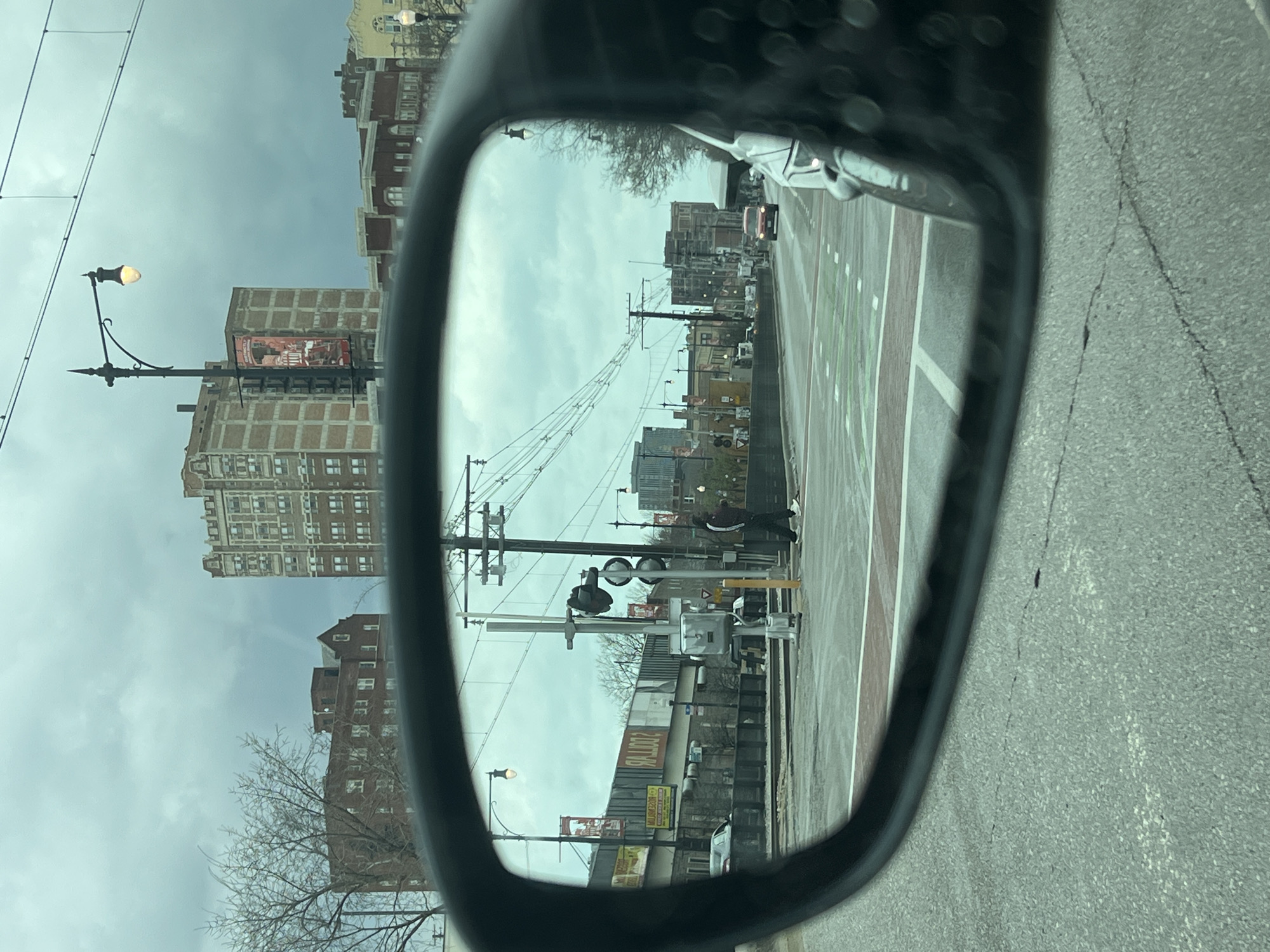 The image shows a view in a car side mirror, reflecting a street scene. The street features buildings, light posts, power lines, and a pedestrian crossing with railroad crossing signals. A person is walking on the crosswalk. Commercial signage is visible on some buildings. Based on the architecture and street layout, the location is likely in Chicago, Illinois, USA, but specific landmarks are not clearly identifiable in the reflection to pinpoint the exact address. The weather appears overcast. The road next to the car is cracked and gray.