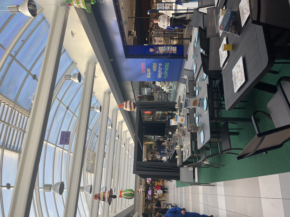 The image shows a view of an indoor shopping mall in Saint-Brice-Courcelles, France. The image shows a large, open-air shopping mall, featuring an open-plan design with a glass roof. There are several shops on either side of the mall, with a central walkway paved in black and white tiles. On one side of the walkway, a display of outdoor furniture is set up. This display consists of four black tables, six gray chairs, and a green rug. There are also several price tags attached to the tables. The space is decorated with festive Christmas decorations, including bells hanging from the ceiling. The mall is lit by multiple overhead lights. On the opposite side of the walkway, people can be seen walking, and several shops can be seen, one of which appears to be a multimedia store. A man is seen talking to a cashier. There is a sign in the store window that reads "Librairie, Disques, Video, Telephonie, Multimedia". A glass door leads into the store, revealing a well-lit and stocked interior. The store windows and doorways are guarded by black metal poles. There is a sign reading "Bien" in the distance.