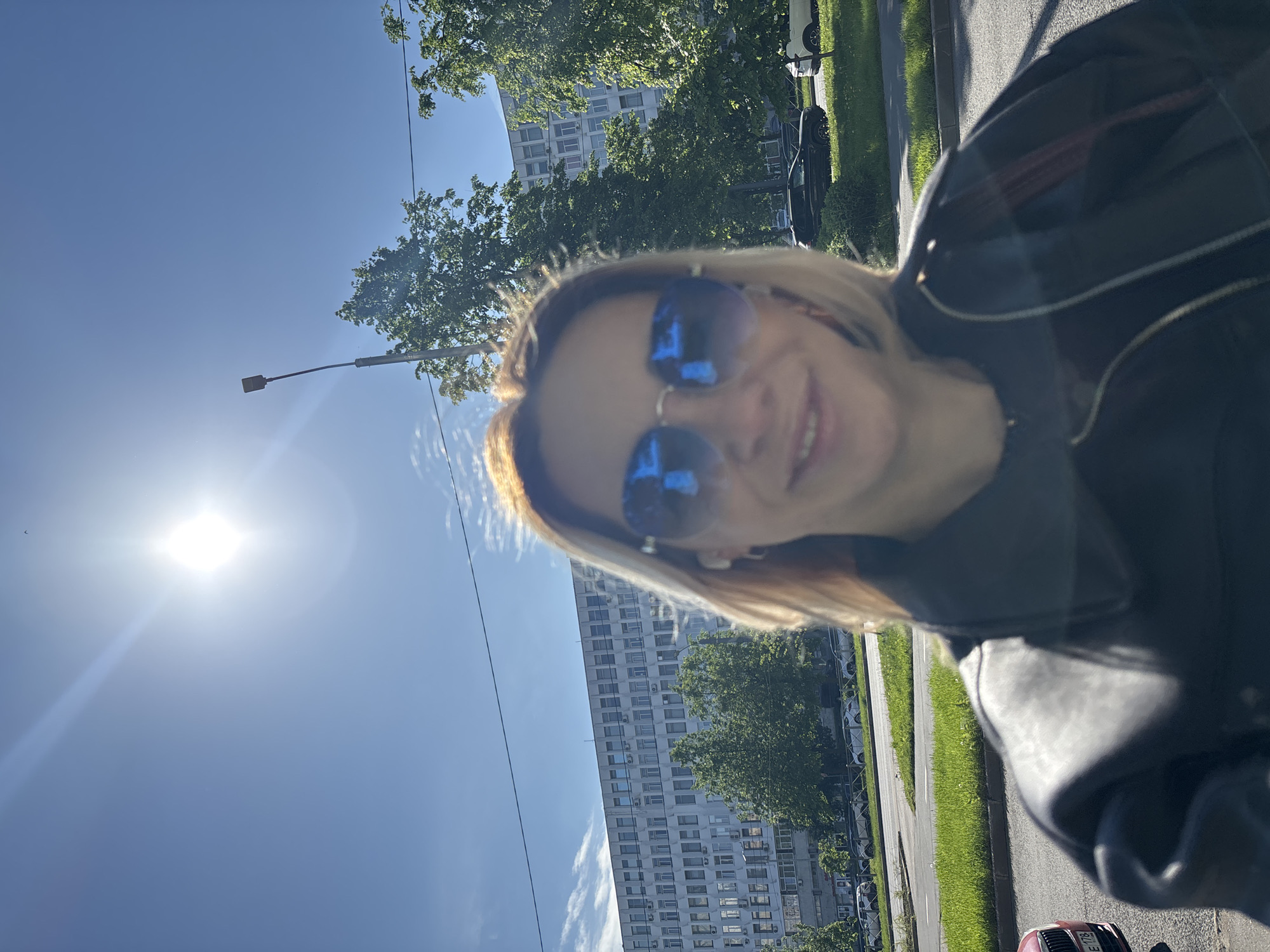 The media file is a bright, sunny outdoor selfie, presented rotated 90 degrees clockwise. The primary subject is a woman, smiling, whose blonde hair is visible around her face. She wears blue reflective sunglasses, which cover her eyes, and a dark jacket, possibly leather, which is visible on her shoulders and part of her arm.
The background reveals an urban or suburban setting under a clear blue sky. A brilliant sun, a strong light source, is positioned in the upper left of the correctly oriented image. To the left, a tall, multi-story building, likely an apartment or office block, featuring many windows and some air conditioning units, stands partially framed by green trees. On the right side, there is a street or road with a paved surface, a curb, and a strip of green grass, beyond which several cars are parked in the distance. A lamppost extends into the frame from the mid-left.
The time of day is bright, likely morning or afternoon, given the high sun. The clear sky indicates good weather. The woman's expression conveys a positive and happy mood. A small red section of a vehicle, possibly a car or motorcycle, is visible in the bottom right corner of the correctly oriented image, displaying a license plate with the visible numbers "116".