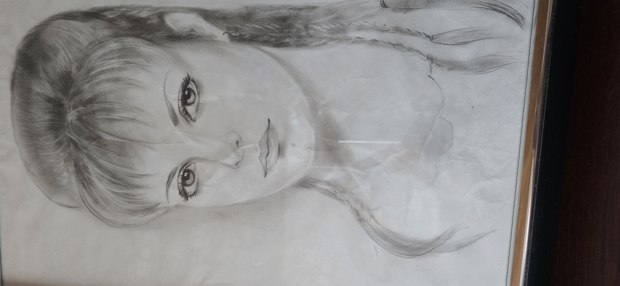 A black and white pencil sketch of a woman's face is framed in a thin silver and black frame. The drawing shows the woman from the shoulders up, with her head slightly turned to the left. She has dark, voluminous hair styled in a high updo with bangs. Her eyes are large and expressive, with visible eyelashes. She has a small nose and full lips. Two braids hang down on either side of her shoulders. The sketch is detailed, with shading used to create depth and dimension. The paper appears slightly creased and aged. The background is plain, with no discernible details. The frame rests on a dark wooden surface, possibly a table or shelf. There is no indication of a specific location in Bad Marienberg, Germany, within the image itself.