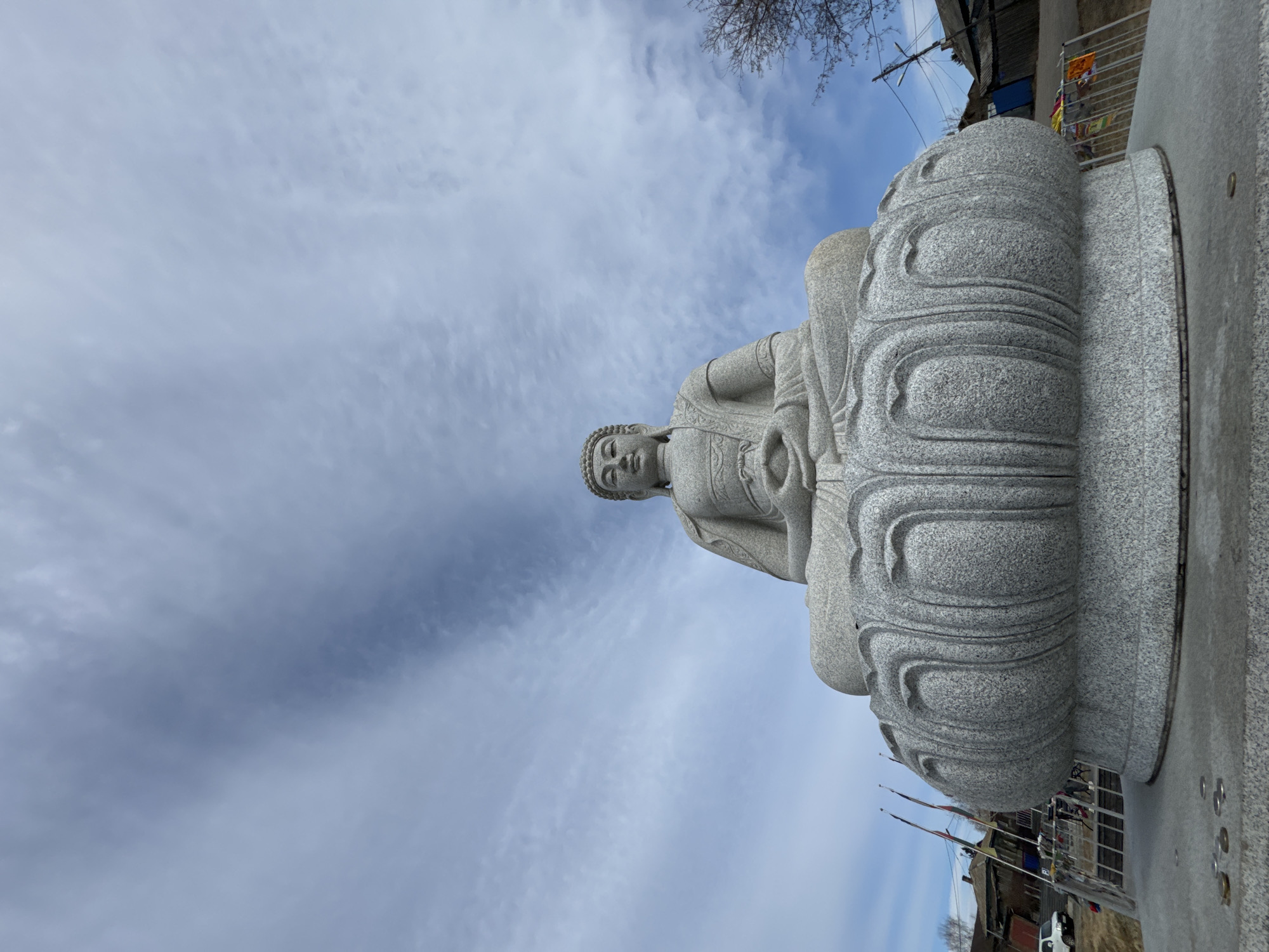 A low-angle, outdoor, daytime shot depicts a gray stone Buddha statue in the city of Krasnoyarsk, Russia. The statue is seated on a lotus-shaped pedestal. The sky is partially covered with light, wispy clouds. The background includes buildings, trees, flags, and vehicles.