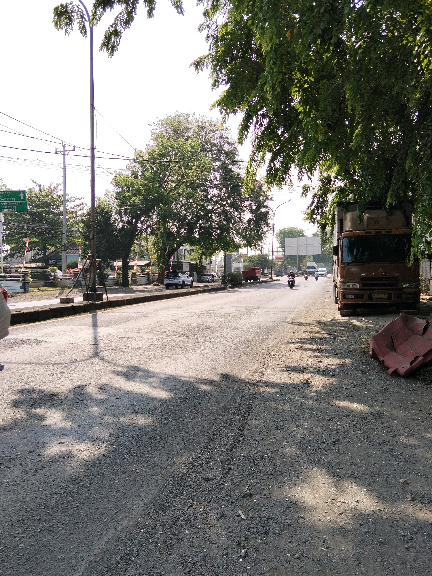 The image depicts a roadside scene in Kasepuhan, Indonesia, under bright daylight with distinct shadows cast by trees and overhead wires. The foreground features a dusty, gravelly shoulder bordering an asphalt road. On the right, a large, rusty brown truck is parked partially on the shoulder, facing the road. Next to it, a red plastic barrier lies on the ground.
The road itself is moderately busy with traffic. Several motorcycles are visible, with one prominently in the middle ground to the left of the truck, and another further down the road. A white van and a larger truck are also seen traveling in the distance on the road.
To the left of the road, a sidewalk and a low wall are present. Behind the wall, a cluster of buildings with red and white flags can be observed. A prominent green directional sign is also visible on the left, indicating directions to "Batang", "Kendal", "Semarang", and "GOR Sarengat". A tall lamp post stands near the sidewalk, with its shadow stretching across the road. Green trees with lush foliage line both sides of the road, with some branches extending over the scene, filtering the sunlight and creating dappled shadows on the ground. The sky is bright and appears clear.