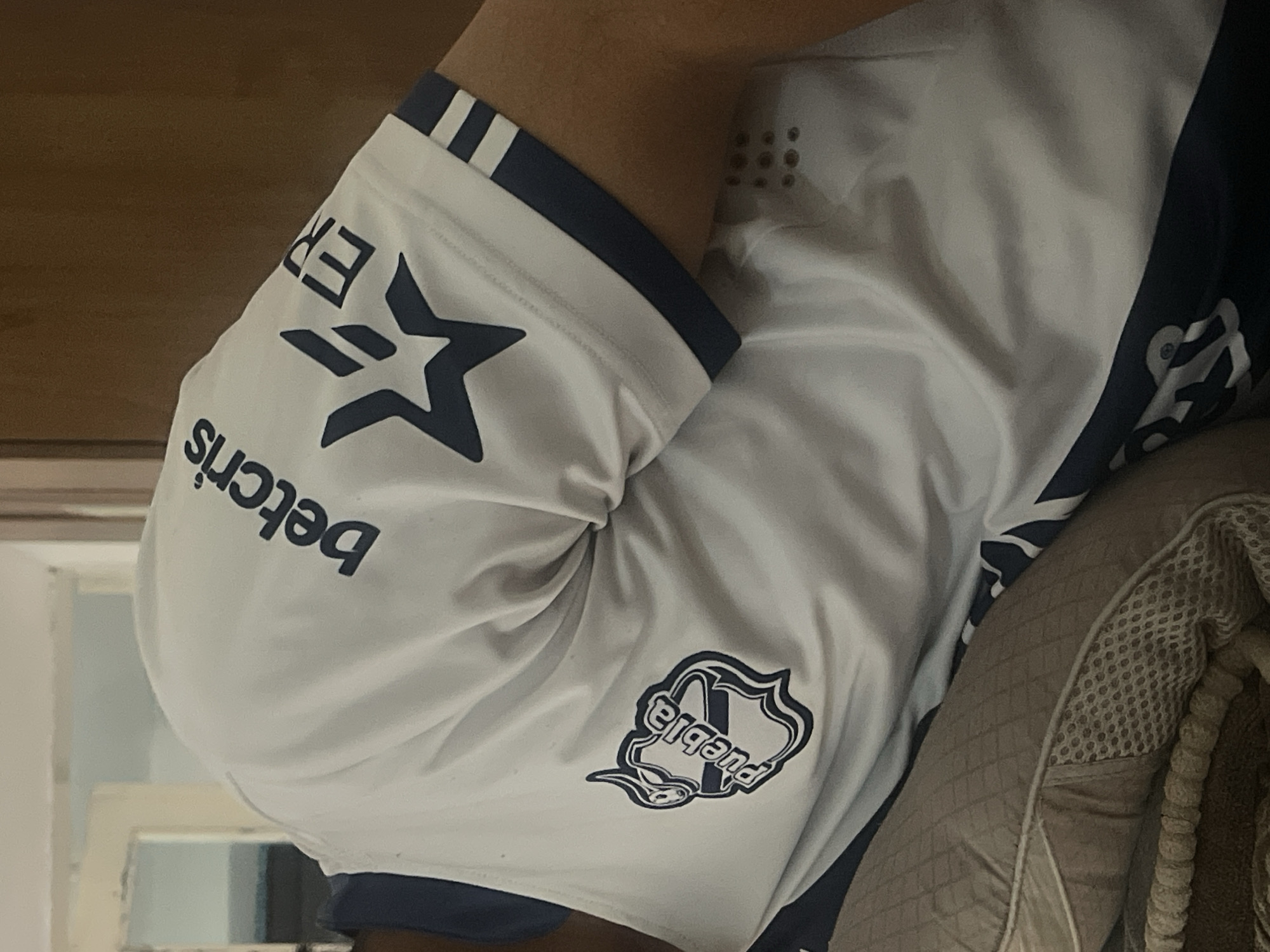 A person is lying down on a bed or couch, wearing a white t-shirt with blue accents. The t-shirt has several logos and text on it, including "betcris" and a star emblem. The person's arm is visible, and they are positioned such that their back is to the viewer. The background is out of focus, but appears to be an indoor setting with some furniture. There is no indication that this scene is located in Bishkek City, Kyrgyzstan.
