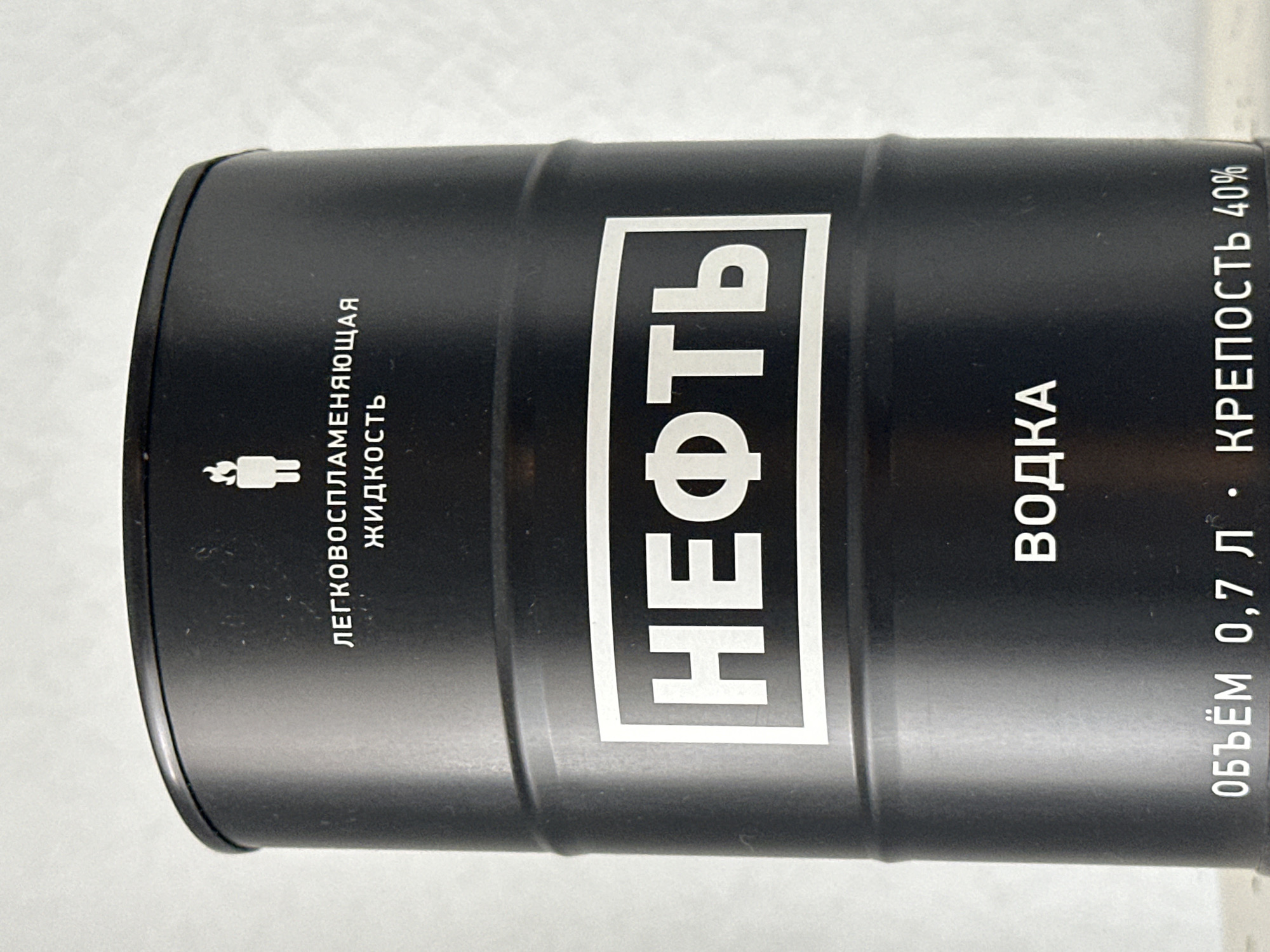 A black, barrel-shaped bottle of vodka stands upright against a light-colored, textured background. The bottle features white text and graphics. Prominently displayed in the center is the word "НЕФТЬ" (oil) in large, bold, white letters within a white rectangle. Above this, smaller white text reads "ЛЕГКОВОСПЛАМЕНЯЮЩАЯСЯ ЖИДКОСТЬ" (flammable liquid), accompanied by a white pictogram of a flame above a human figure. Towards the bottom, the text "ВОДКА" (vodka) appears in white, followed by "ОБЪЁМ 0,7 Л" (volume 0.7 L) and "КРЕПОСТЬ 40%" (strength 40%). The lighting is even, highlighting the texture of the bottle and the background. No geographical information specific to Nevinnomyssk, Russia, is visible in the image.